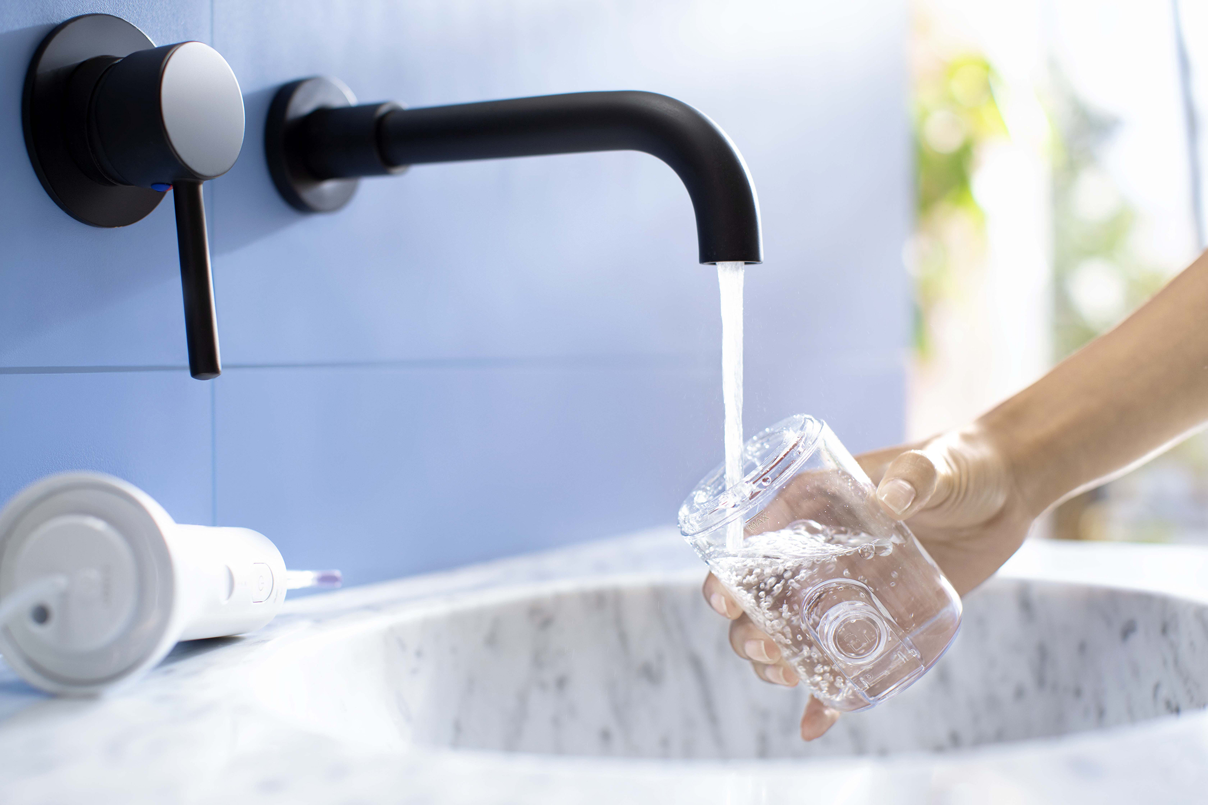 L'eau coule d'un robinet noir dans un verre clair. Un irrigateur buccal blanc est sur l'évier.