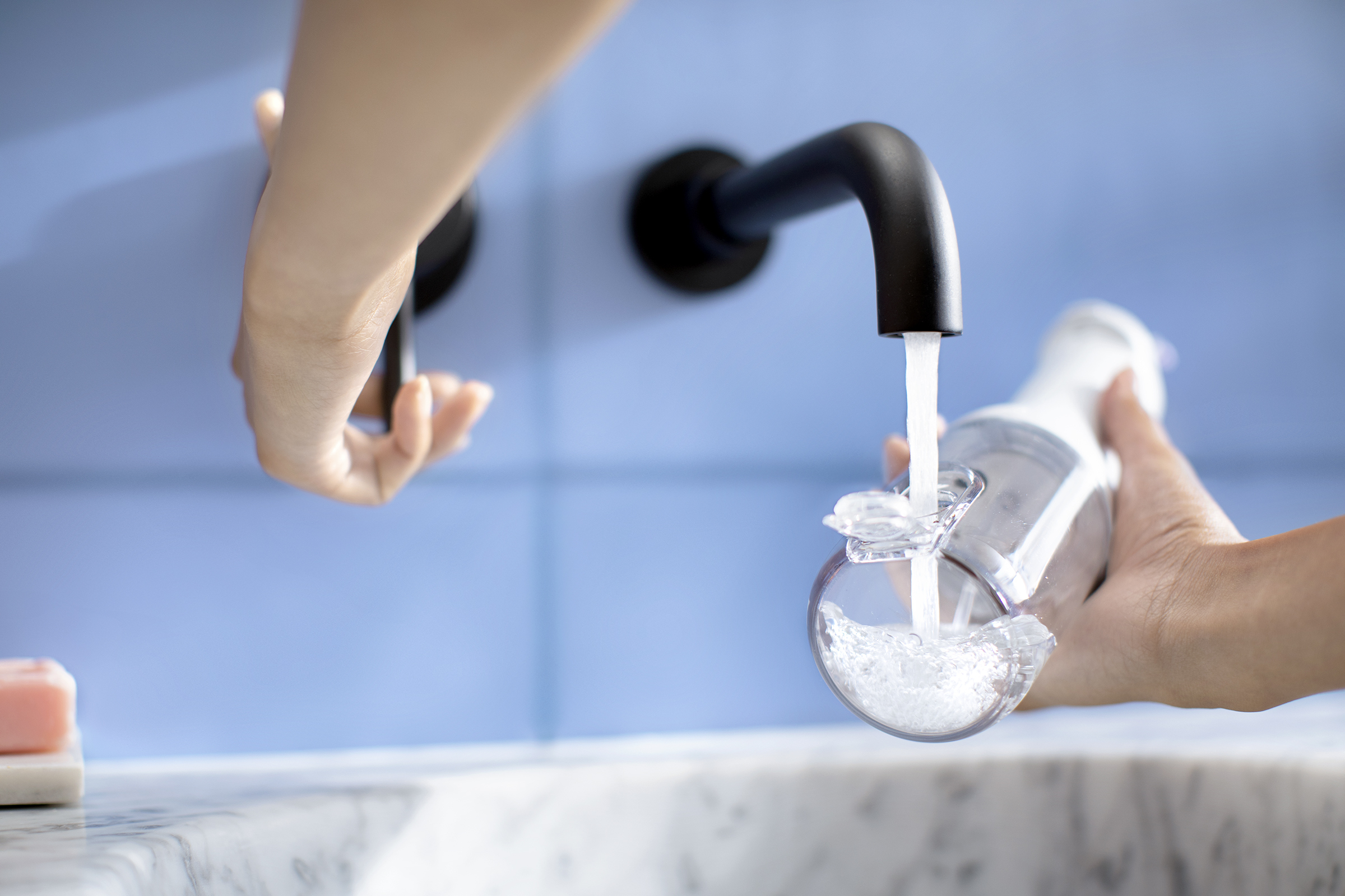 Mains remplissant une bouteille transparente d'eau d'un robinet noir. Fond de carreaux bleus.