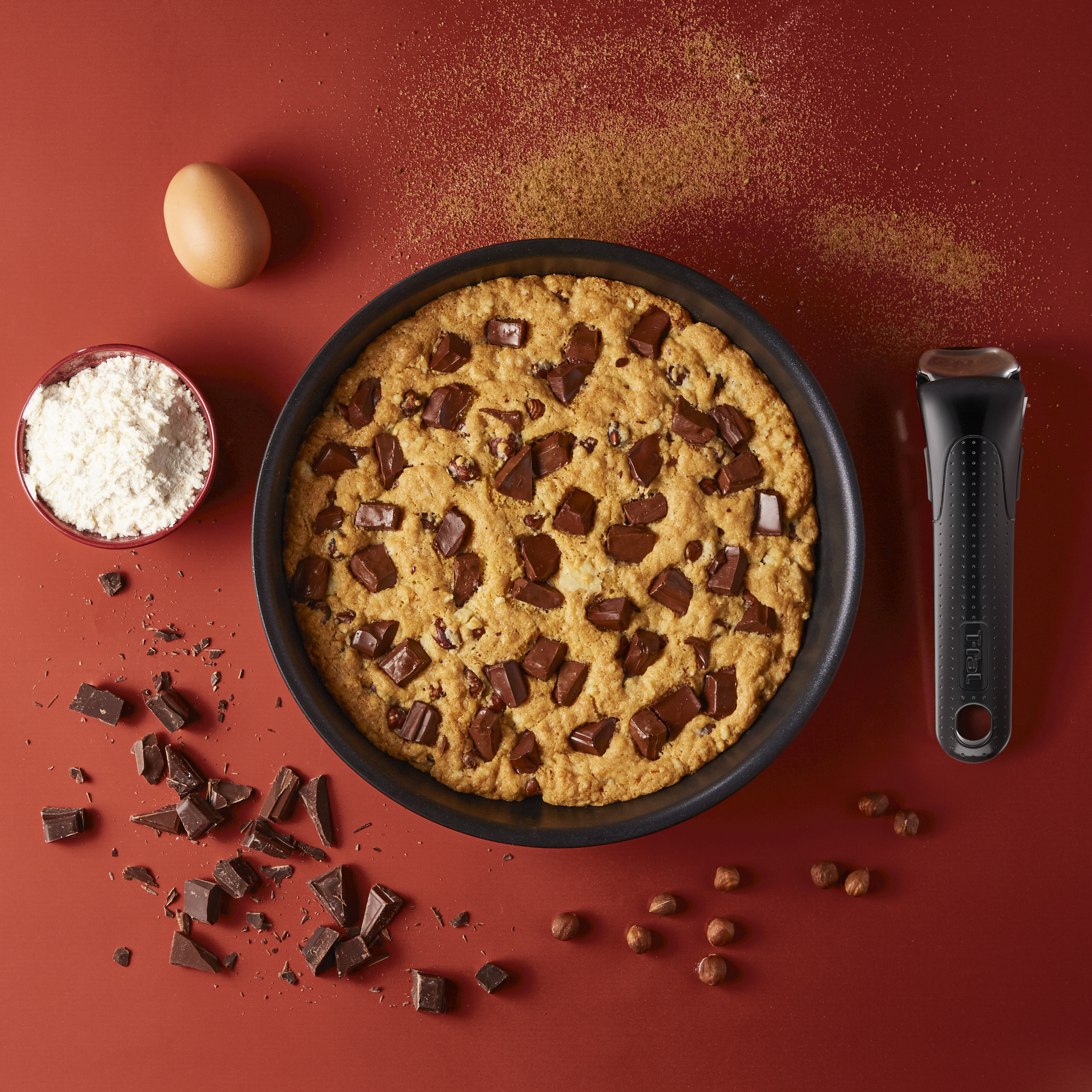 Cookie dans une poêle avec des morceaux de chocolat et des ingrédients. Œuf, farine et une poignée noire sur une surface rouge.