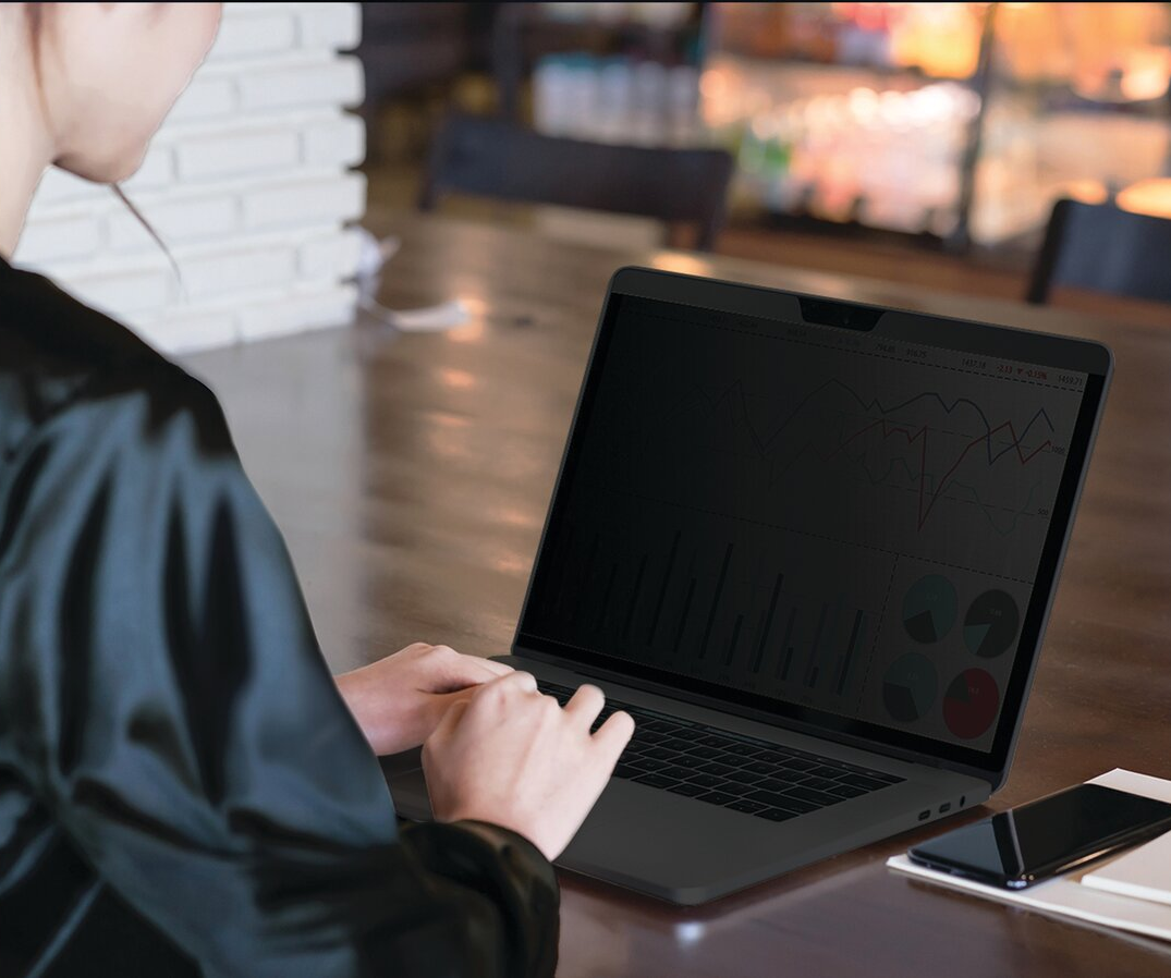 Nő laptopon dolgozik adatvédelmi képernyővel, kávézóban. Diagramok láthatók a képernyőn.
