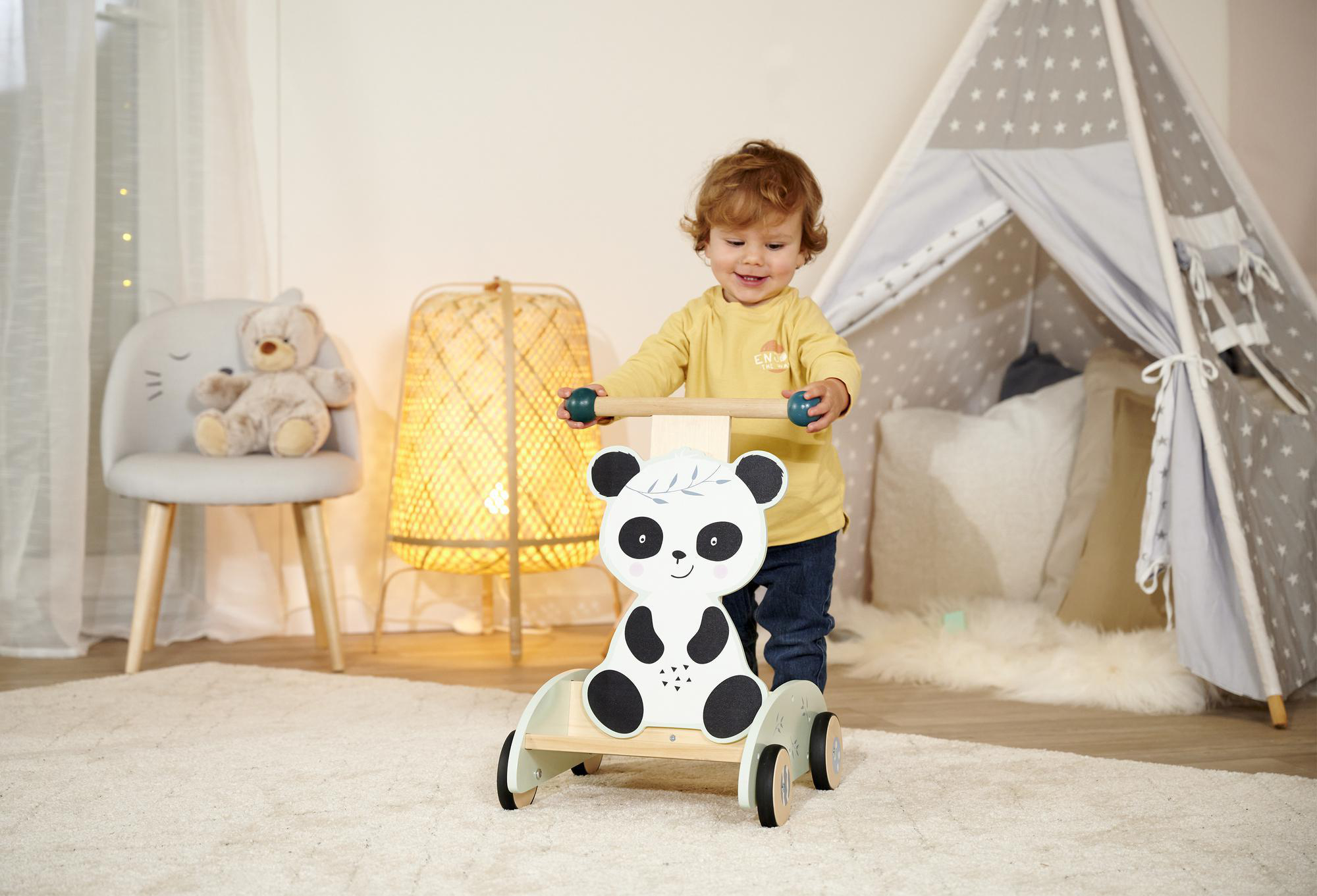 Kleinkind schiebt Panda-Gesicht-Lauflernhilfe auf einem Teppich. Teddybär und Zelt vorhanden.