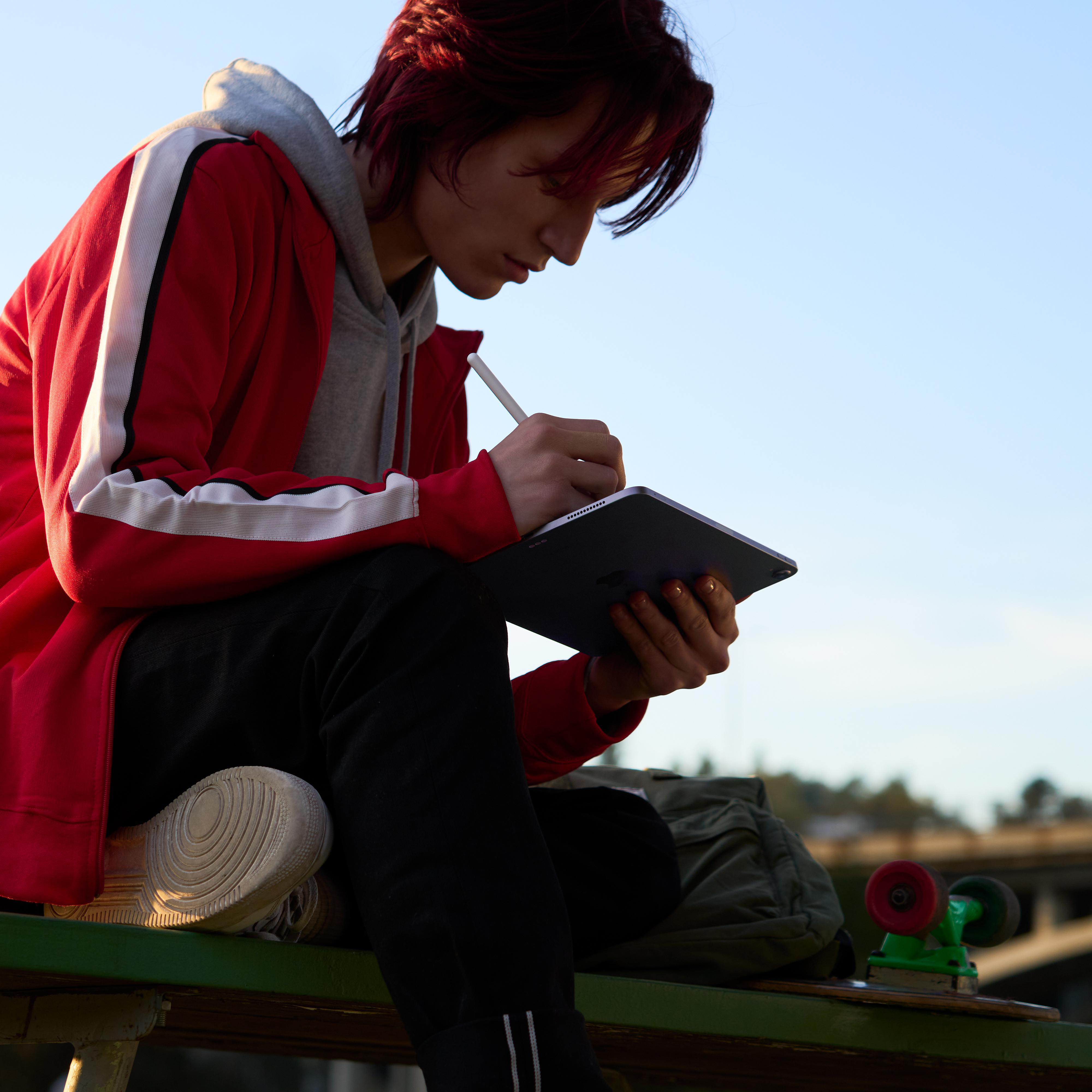 Person schreibt mit Stift auf Tablet, sitzt auf Bank mit Skateboard.