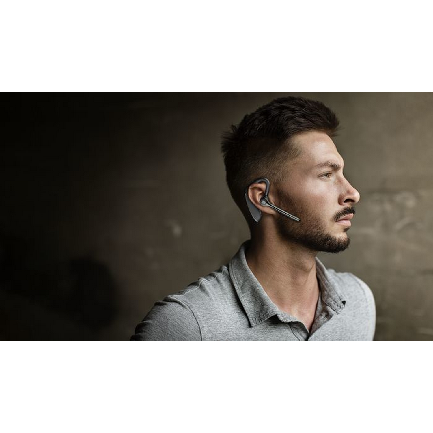 Un hombre que usa unos auriculares Bluetooth negros. Tiene barba y lleva una camisa gris.
