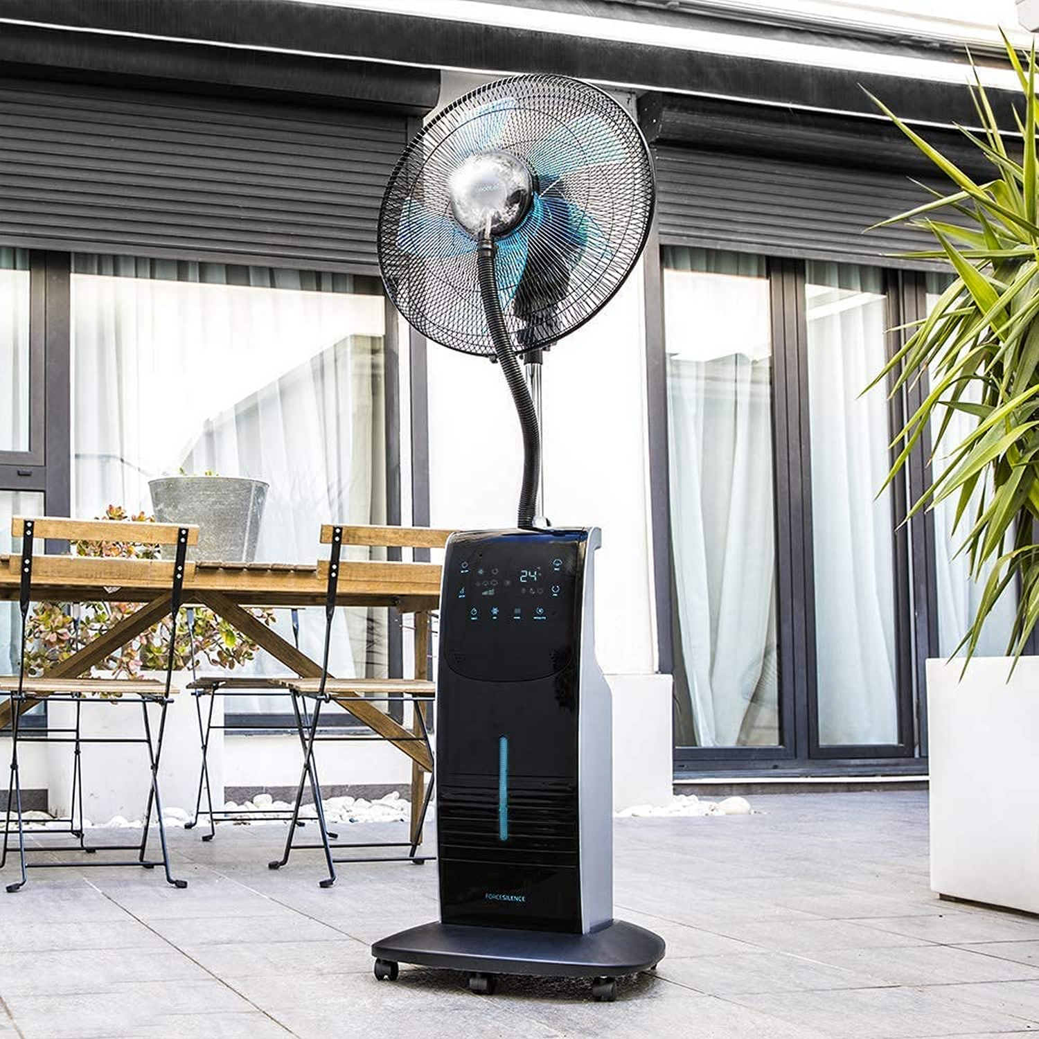 Un ventilador nebulizador negro y gris en un patio. Está junto a una mesa y plantas.