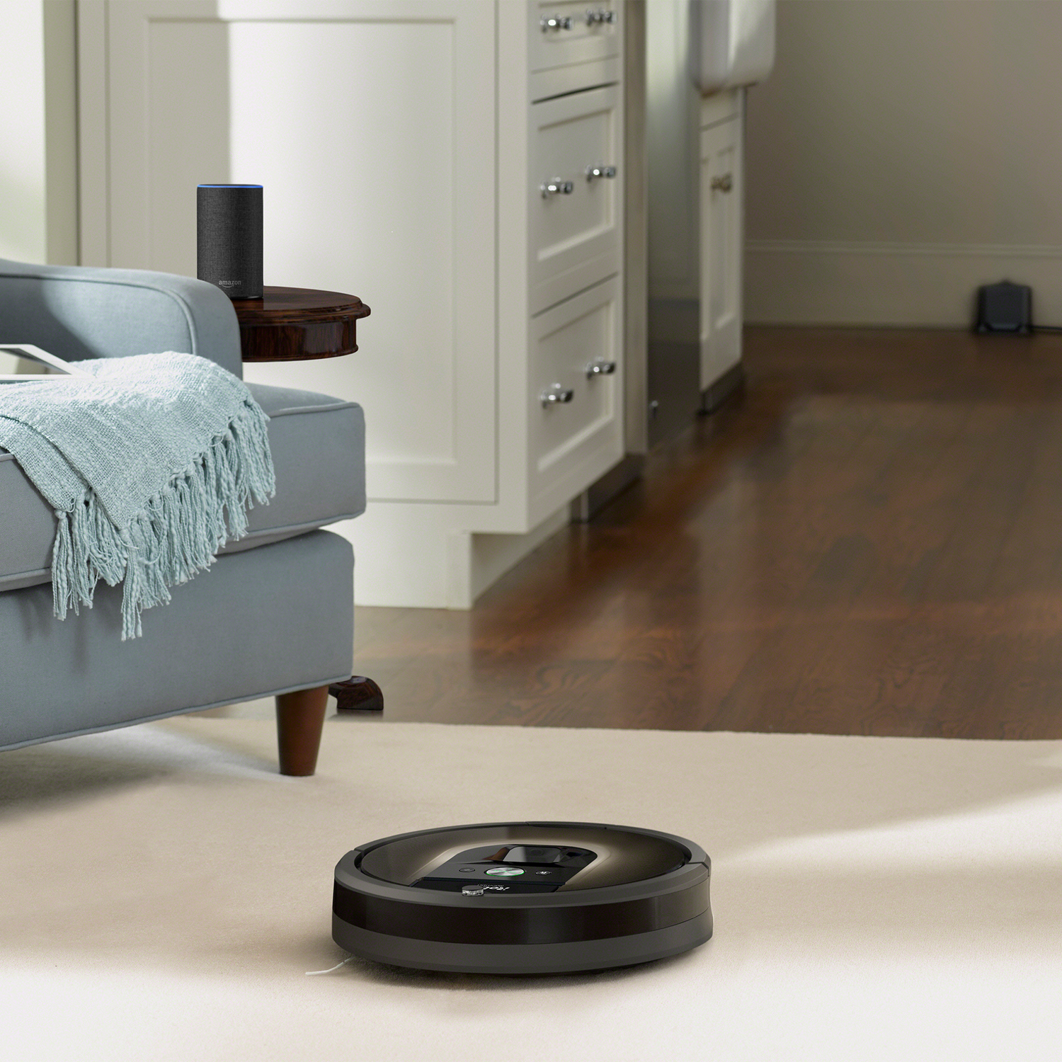 Robot aspirador en una alfombra, con un altavoz Alexa y muebles al fondo.