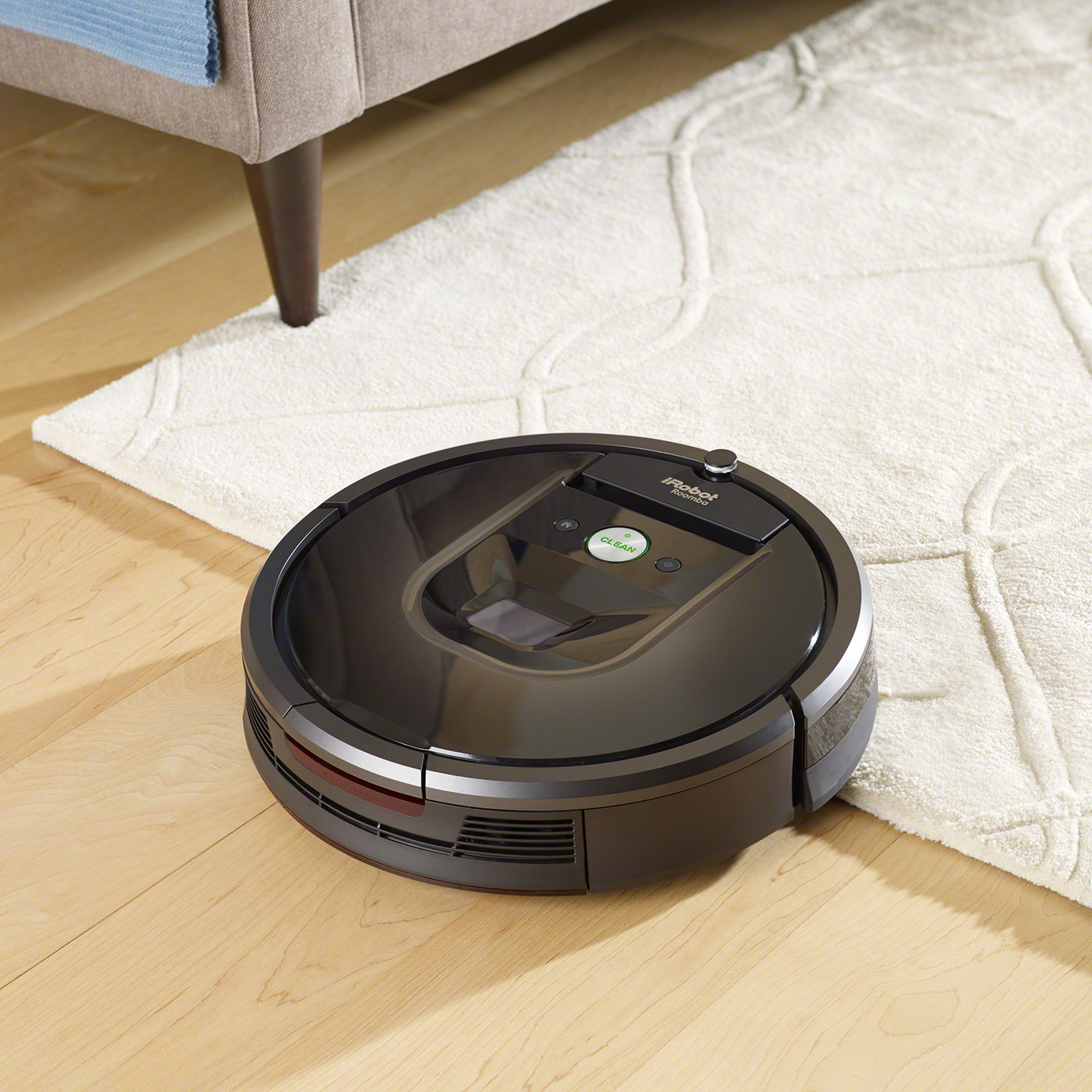 Una aspiradora robot negra en un suelo marrón claro junto a una alfombra blanca.