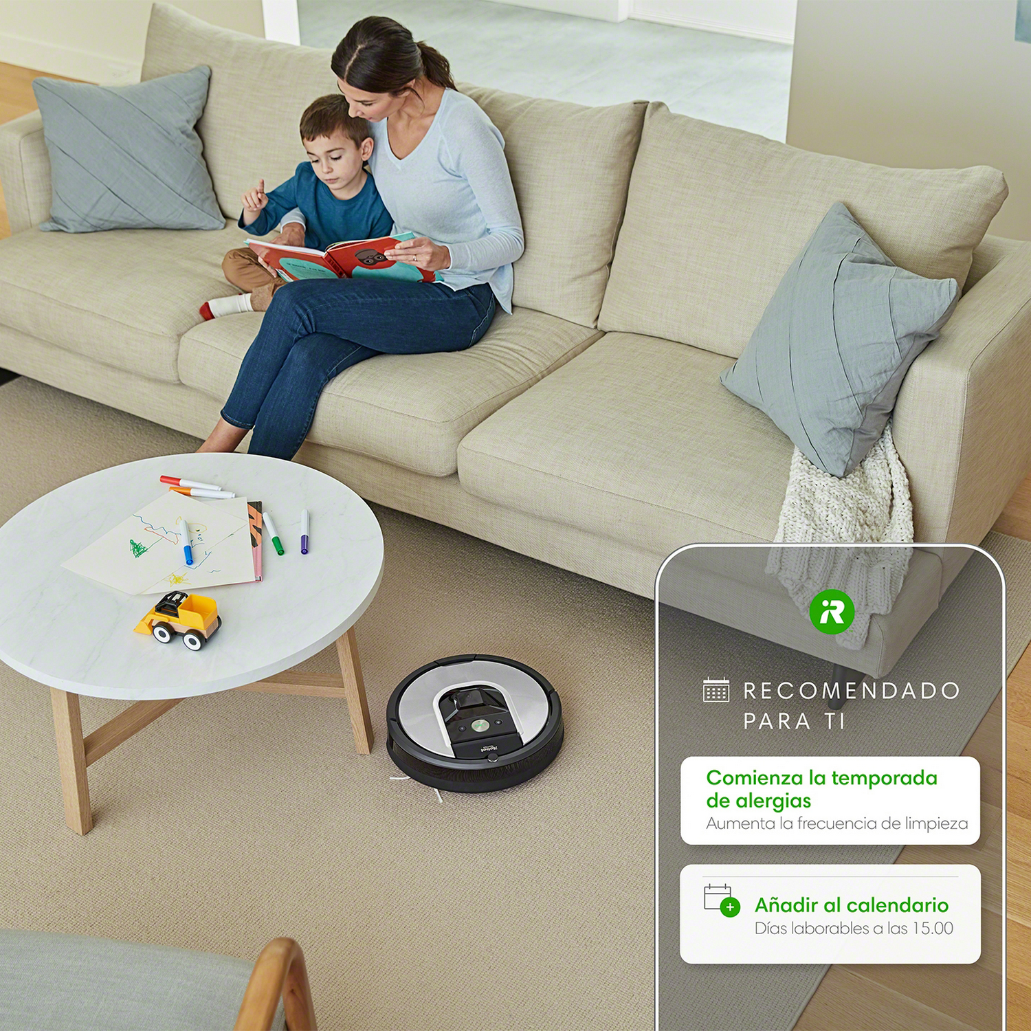Una mujer y un niño leyendo en un sofá con una aspiradora robot y una mesa con útiles de dibujo.