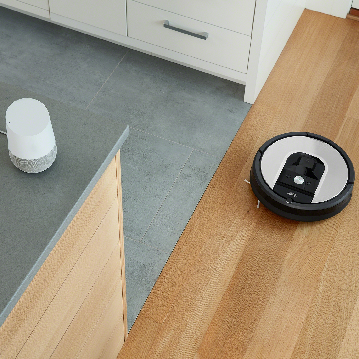Una aspiradora robot y un altavoz inteligente en un piso de madera junto a una encimera.