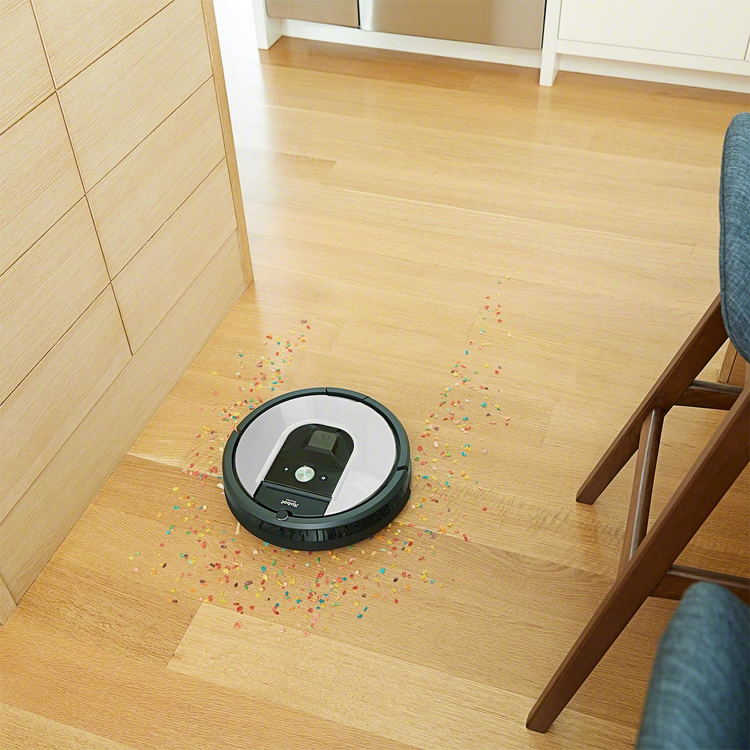 Aspiradora robot en piso de madera, con cereales esparcidos. Silla y gabinetes en el fondo.