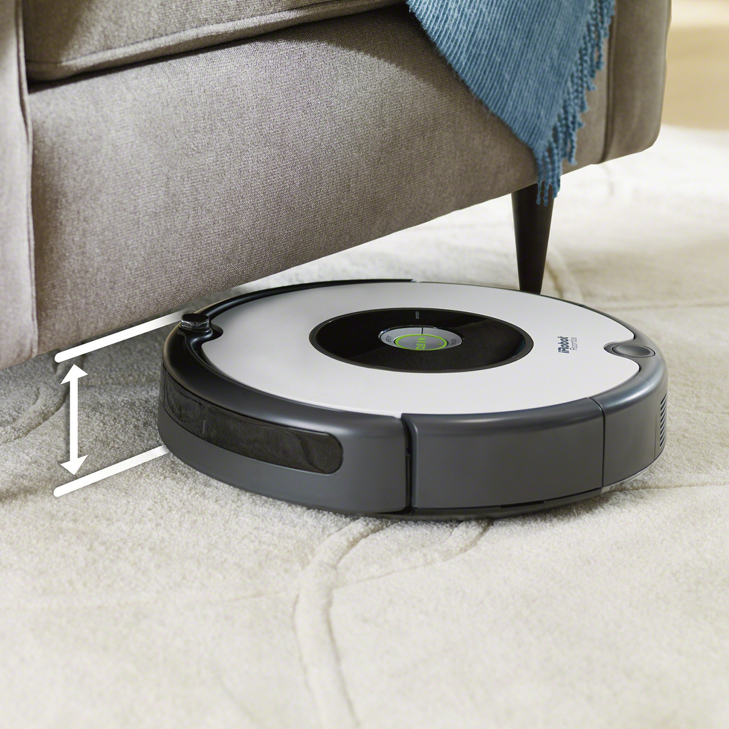 Una aspiradora robot debajo de un sofá, limpiando una alfombra sobre una alfombra blanca.