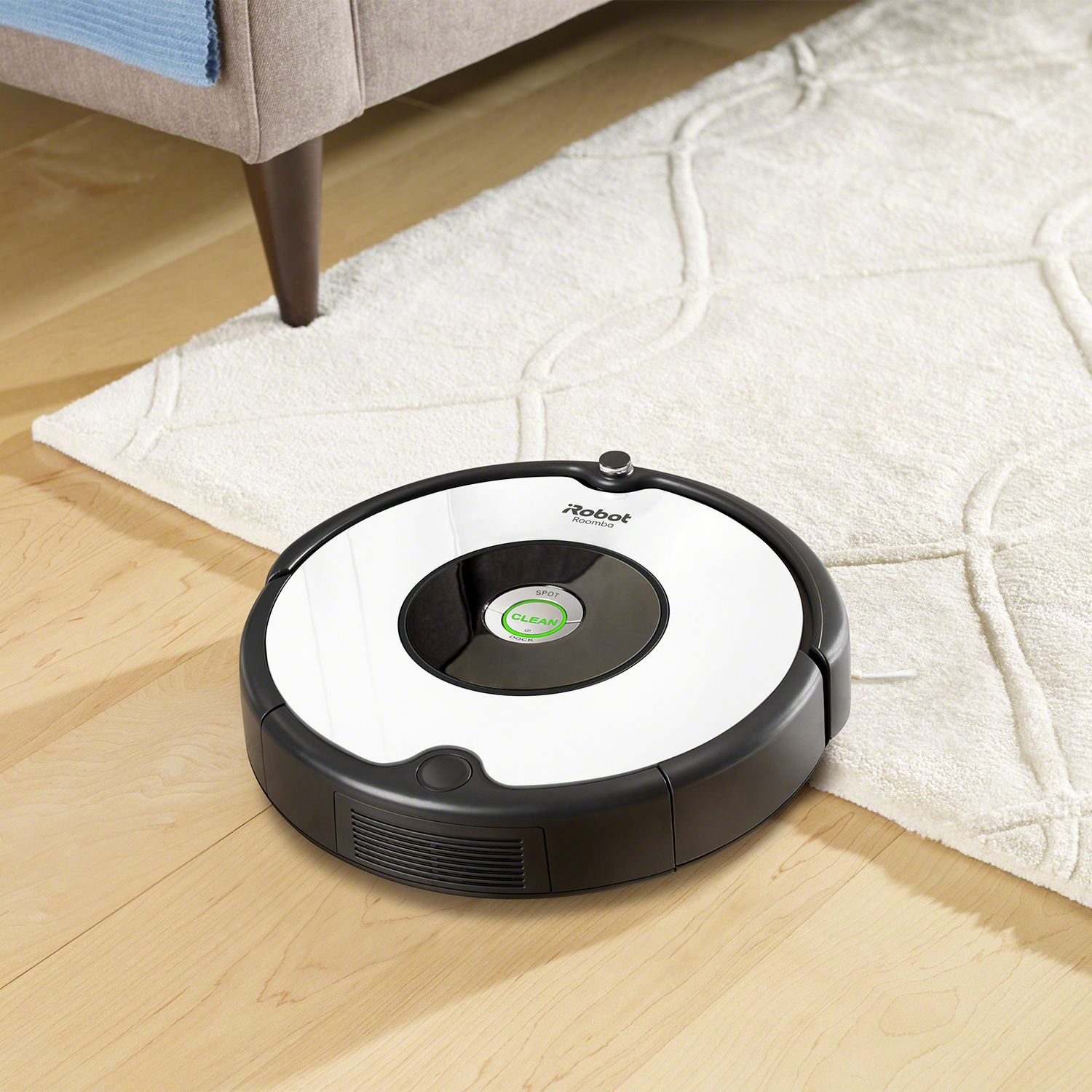 Un robot aspirador blanco y negro sobre un piso de madera, cerca de una alfombra y una pata de mueble.