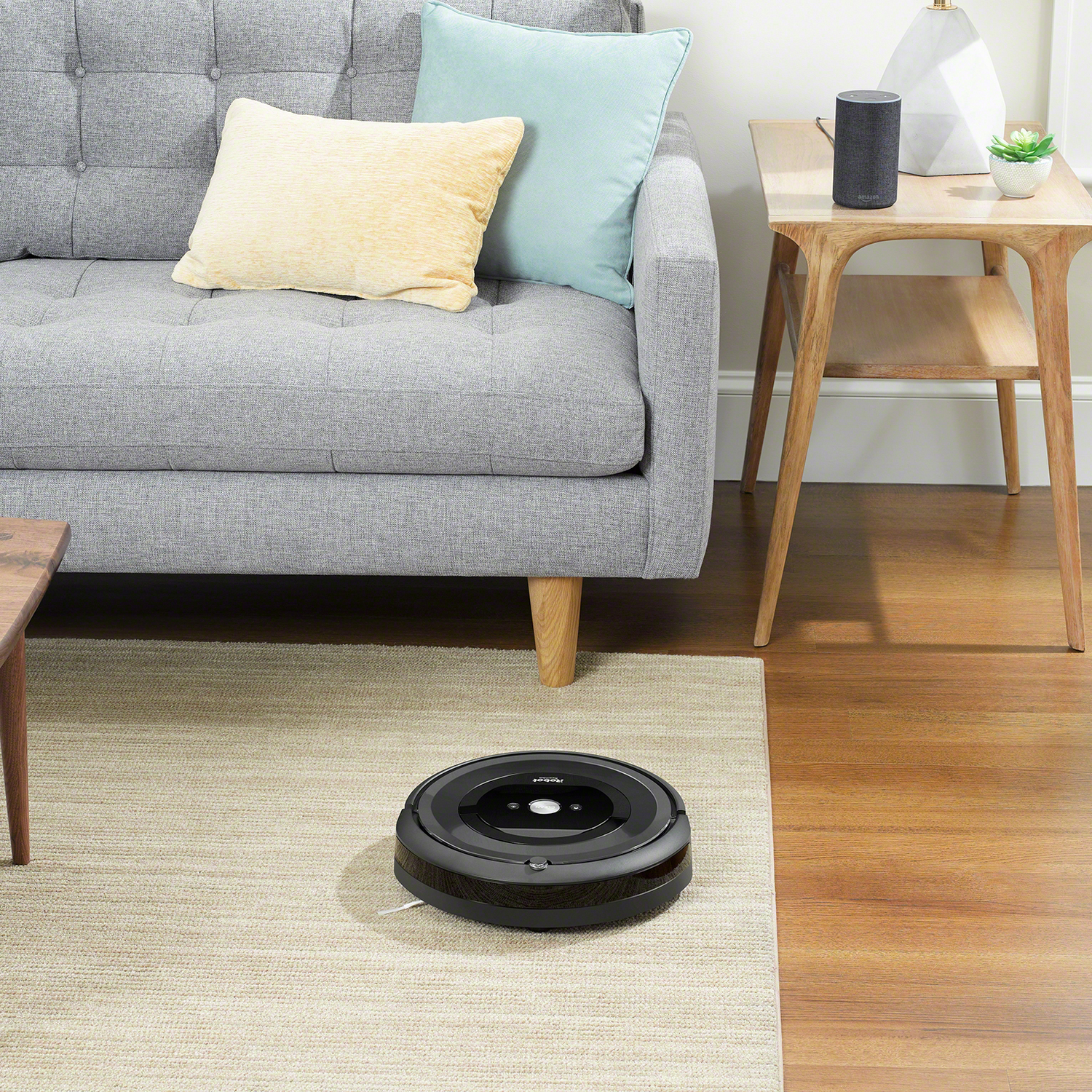 Aspiradora robot negra en una alfombra estampada, cerca de un sofá gris y una mesa auxiliar de madera.