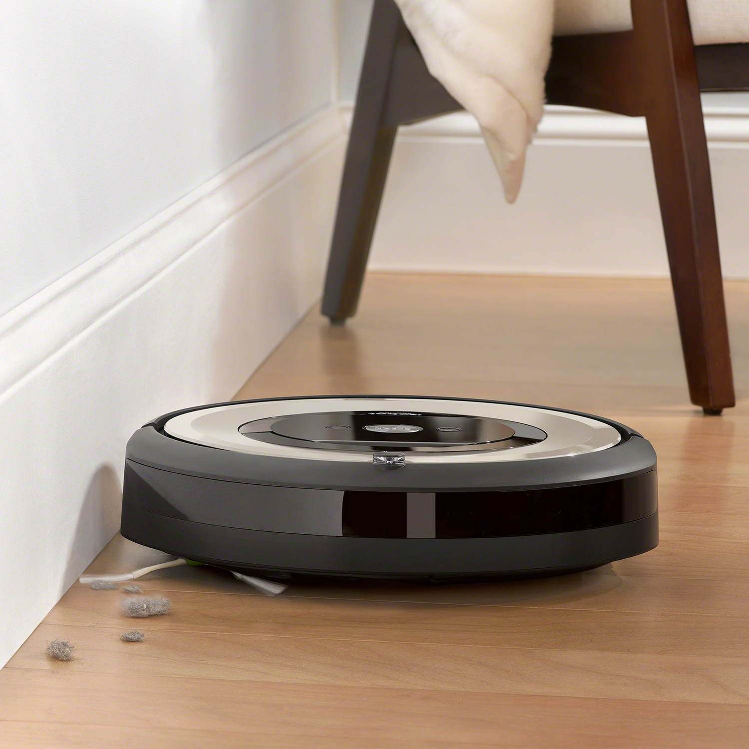 Una aspiradora Roomba limpiando un suelo de madera. Se ve polvo y pelo. La pata de la silla está en el encuadre.