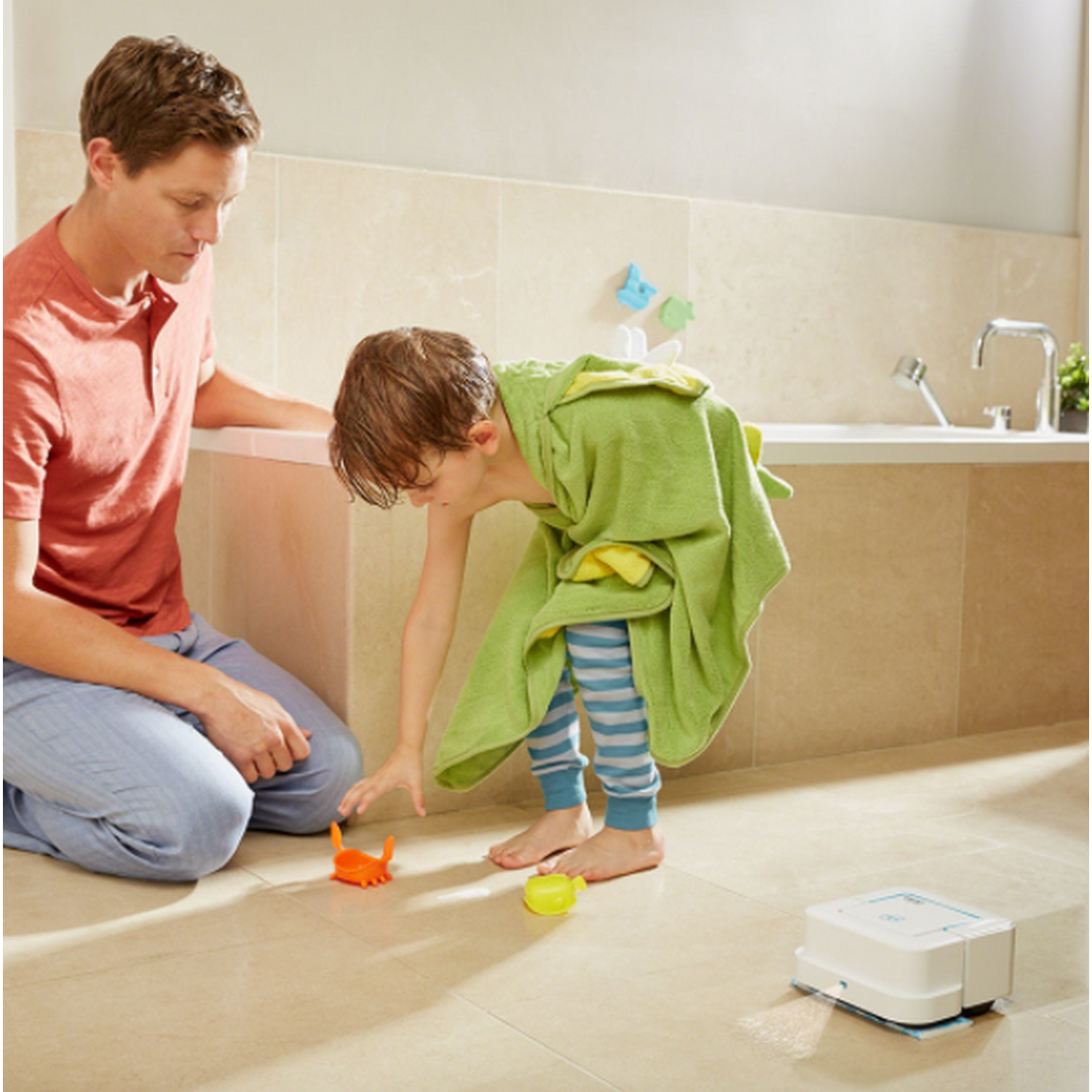 Padre e hijo en el baño. Hijo con toalla verde, jugando con juguetes. Baño blanco, suelo beige.