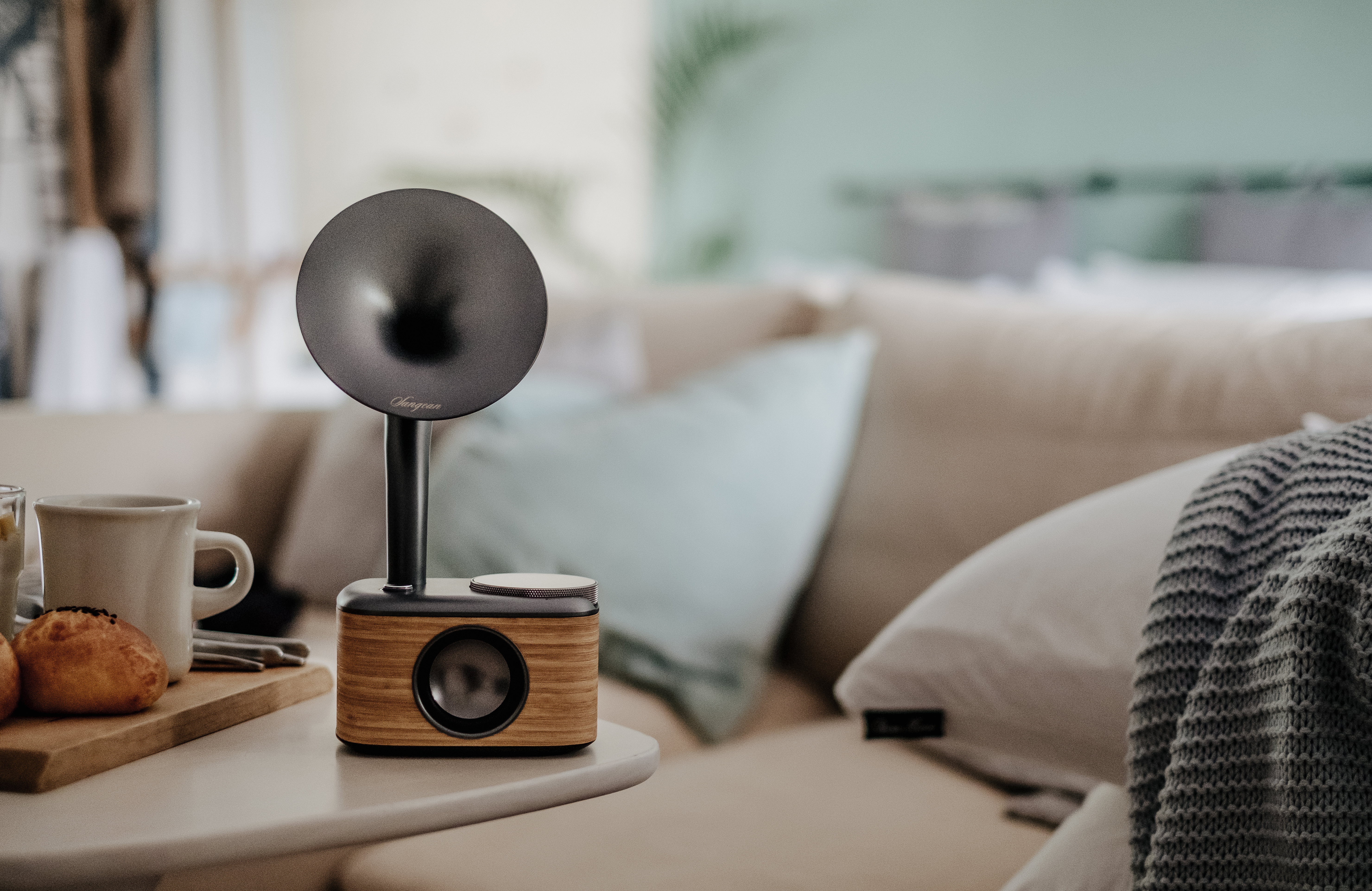 Ein moderner Lautsprecher mit Horn steht auf einem Tisch. Im Hintergrund ein Sofa mit Kissen und einer Decke.