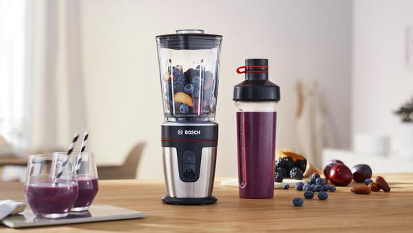 Ein Mixer mit Früchten daneben ein Behälter mit einem lila Smoothie und zwei Gläsern.