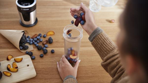 Jemand fügt Blaubeeren und Pflaumen in ein Glas hinzu. Andere Früchte und Mixer sichtbar.