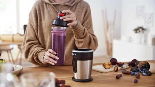 Person, die einen Smoothie zubereitet. Lila Flüssigkeit in einer Flasche. Mixer und Obst auf dem Tisch.