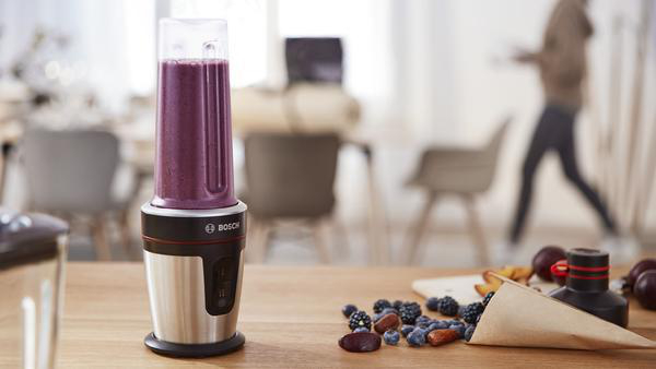 Ein Bosch-Mixer mit einem lila Smoothie, neben Früchten auf einem Holztisch und einer Person im Hintergrund.