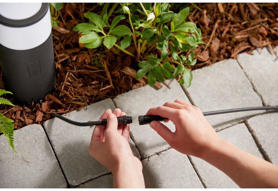 Hands connecting two black cables. A lamp and paving stones are in the background.