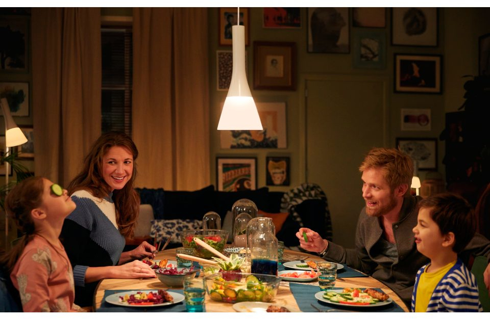 Une famille est assise à une table à manger sous une lampe suspendue blanche, mangeant et parlant.