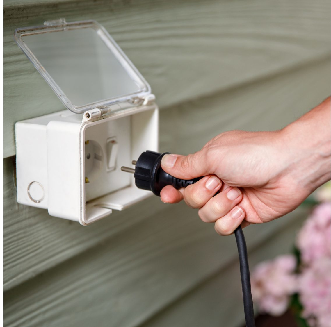 Hand plugging a black plug into an outdoor outlet mounted on a wall.