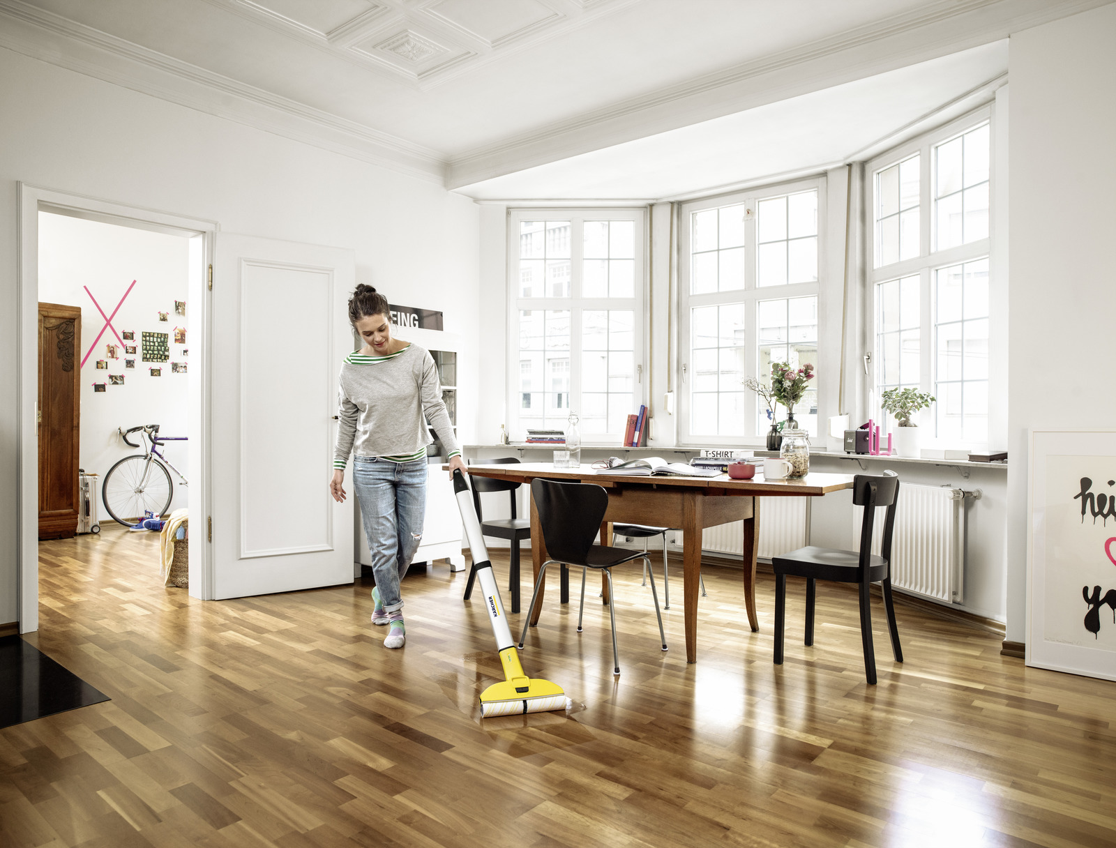 Vrouw die een houten vloer stofzuigt in een lichte kamer. Een fiets en een tafel zijn ook zichtbaar.