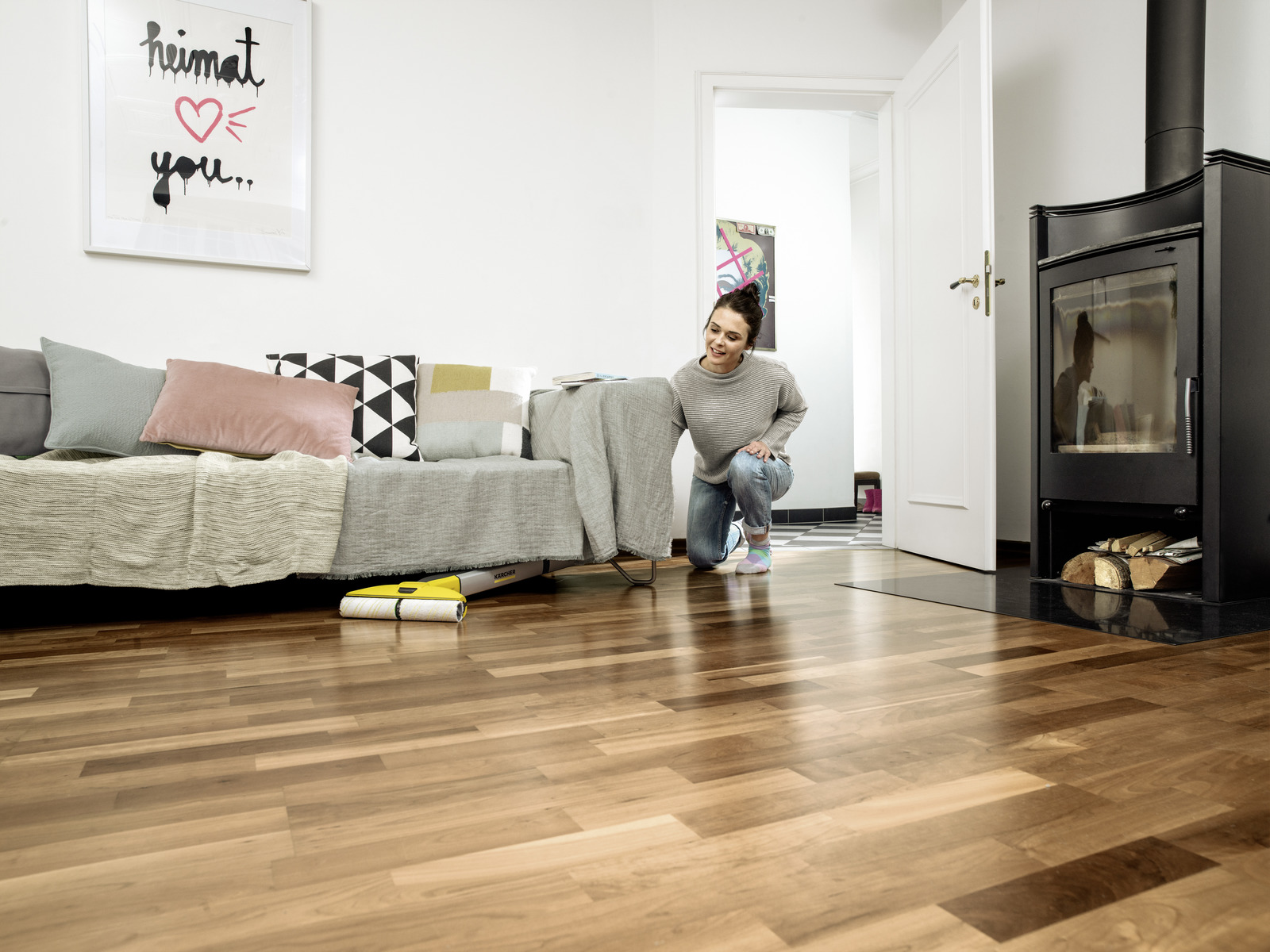 Vrouw knielend bij geel schoonmaakapparaat op houten vloer. Bank, kunstwerk en open haard zichtbaar.