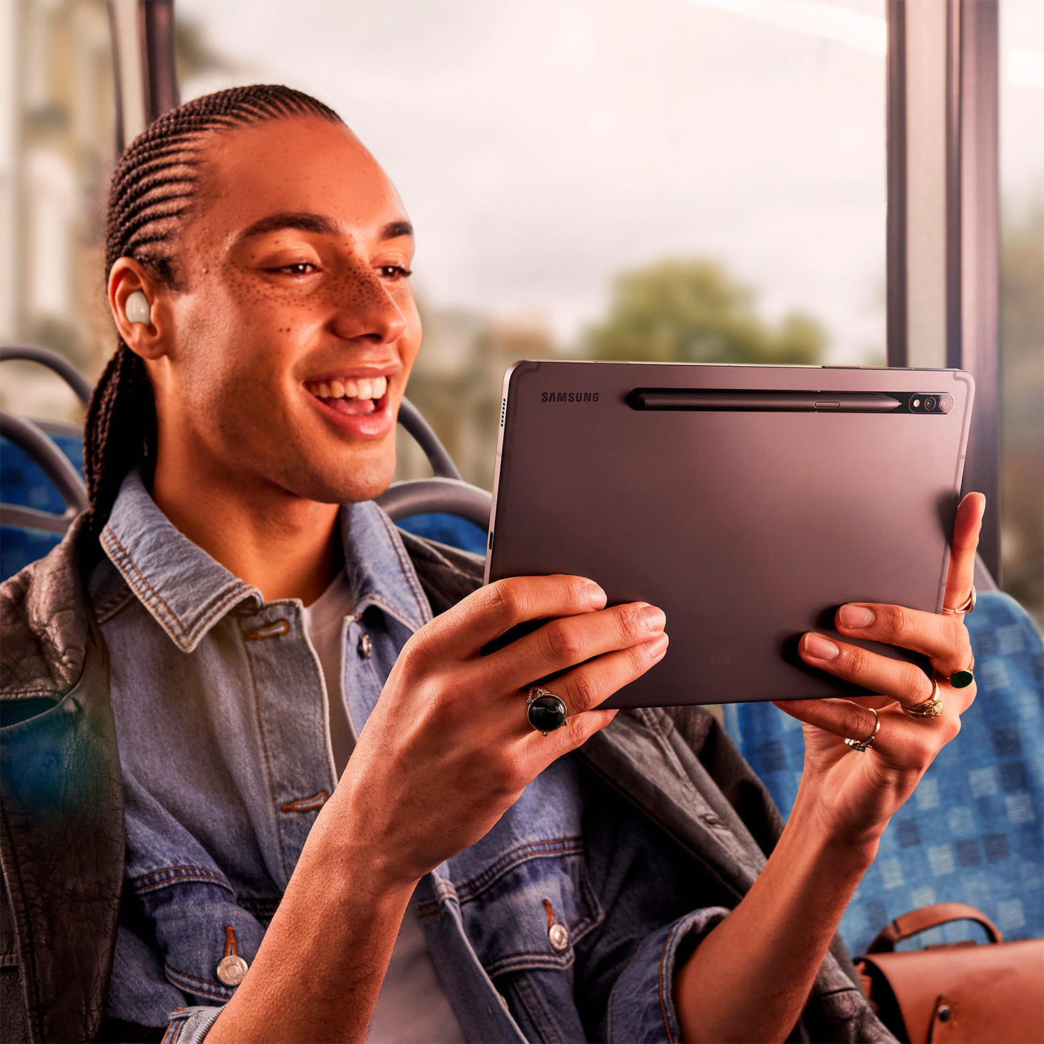 Un hombre sonriente en un autobús, con una chaqueta de mezclilla, usando una tableta. Tiene auriculares en las orejas.