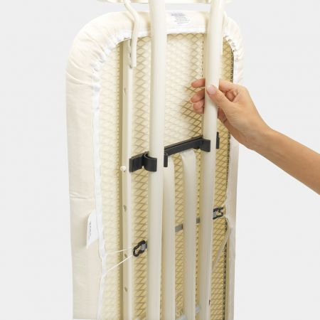 An ironing board, folded. A hand is holding the support. Beige cover and white metal legs.
