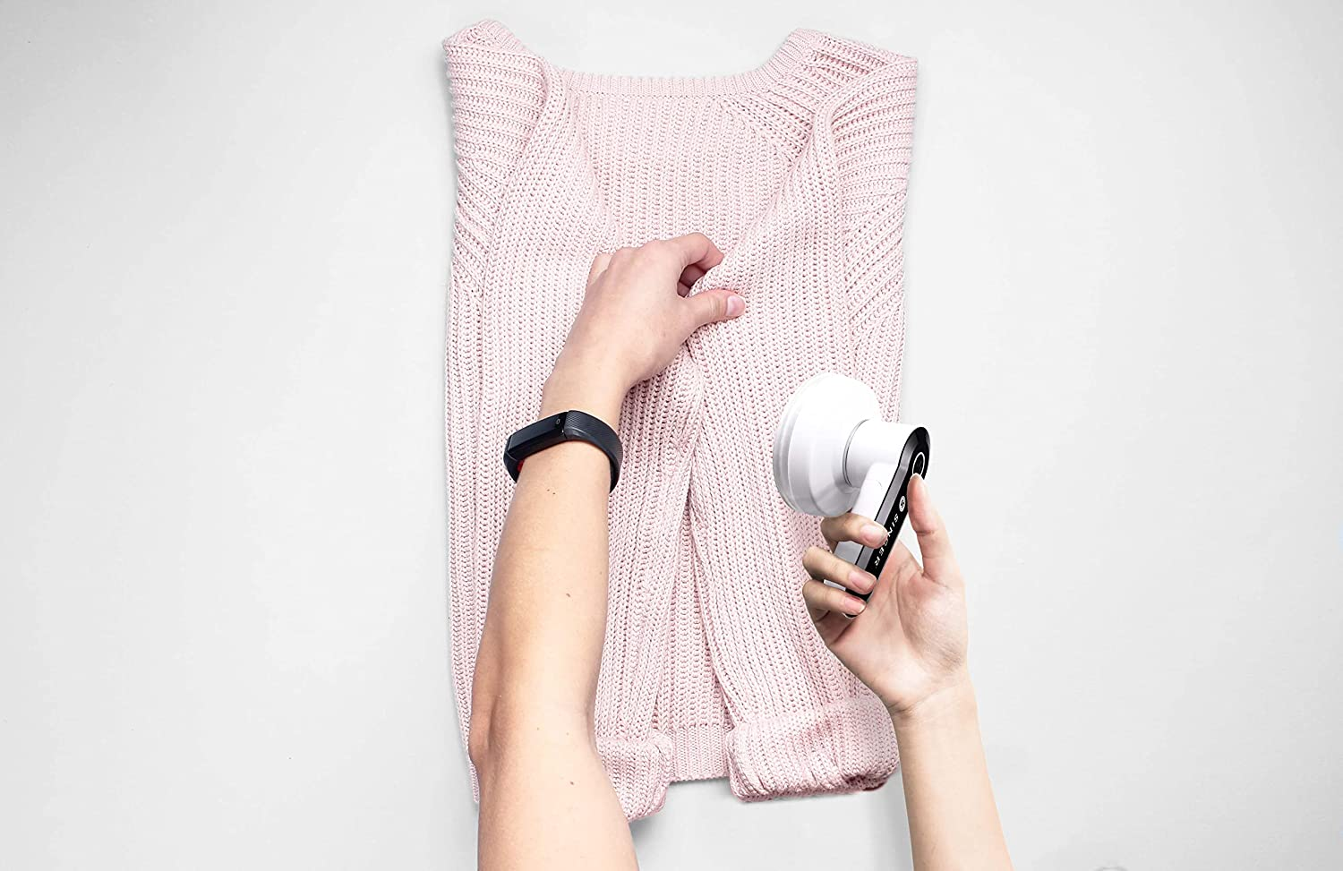 Une personne utilise un anti-bouloches sur un pull rose, posé sur une surface blanche.