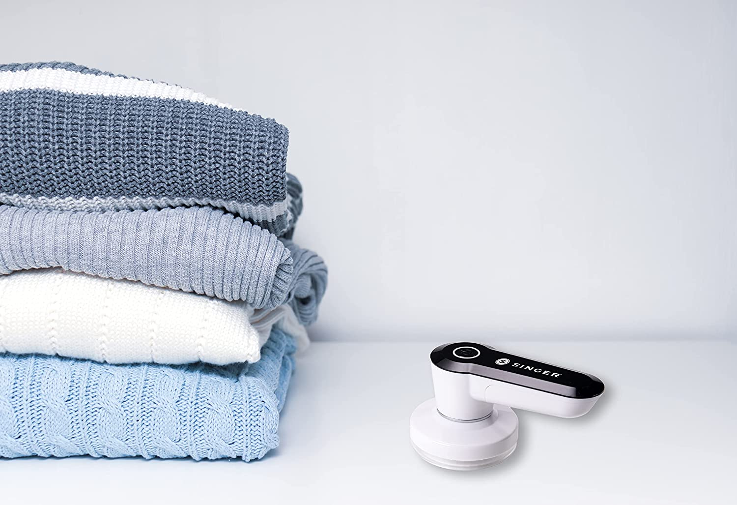 Pile de pulls pliés, rasoir à tissu Singer. Fond blanc.