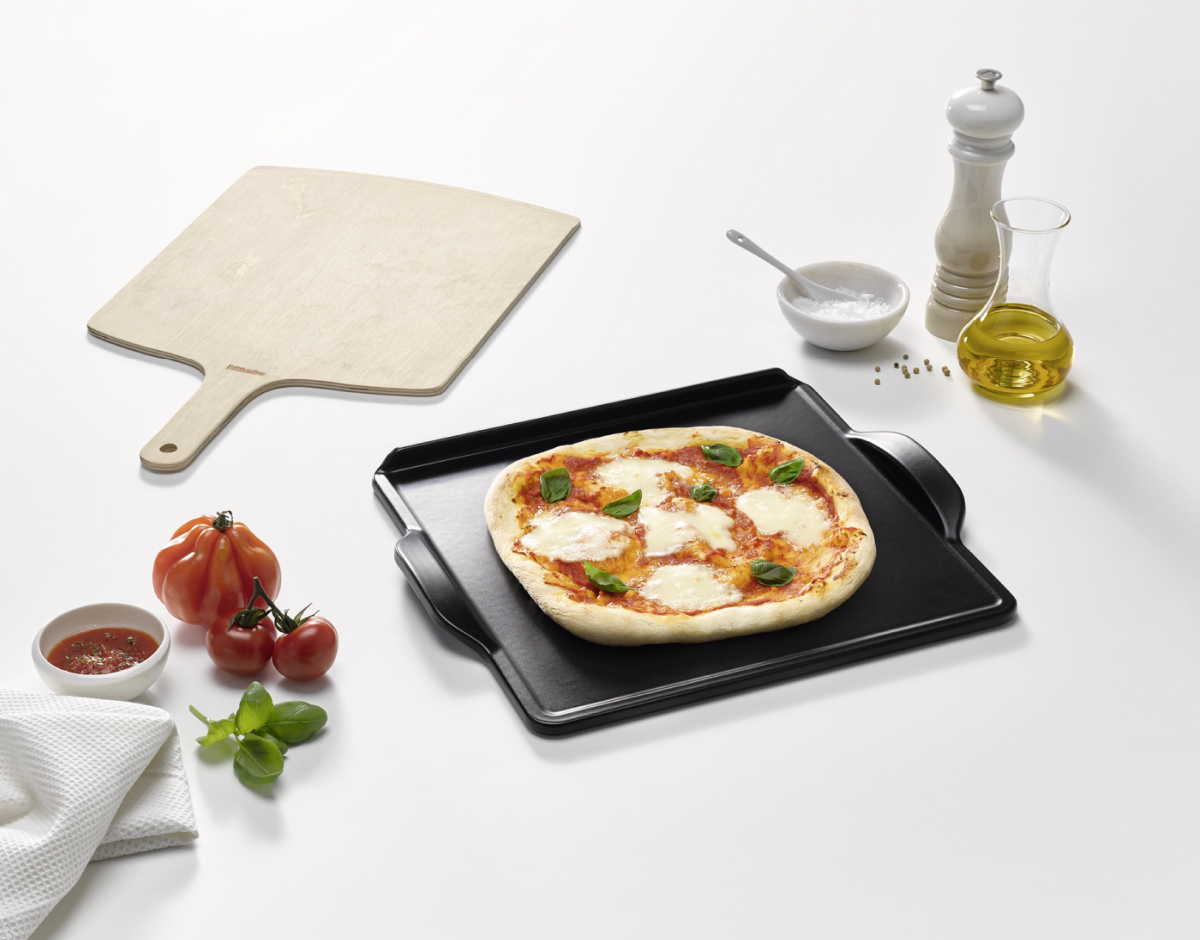 Pizza fekete tálcán, hozzávalókkal, fehér felületen.