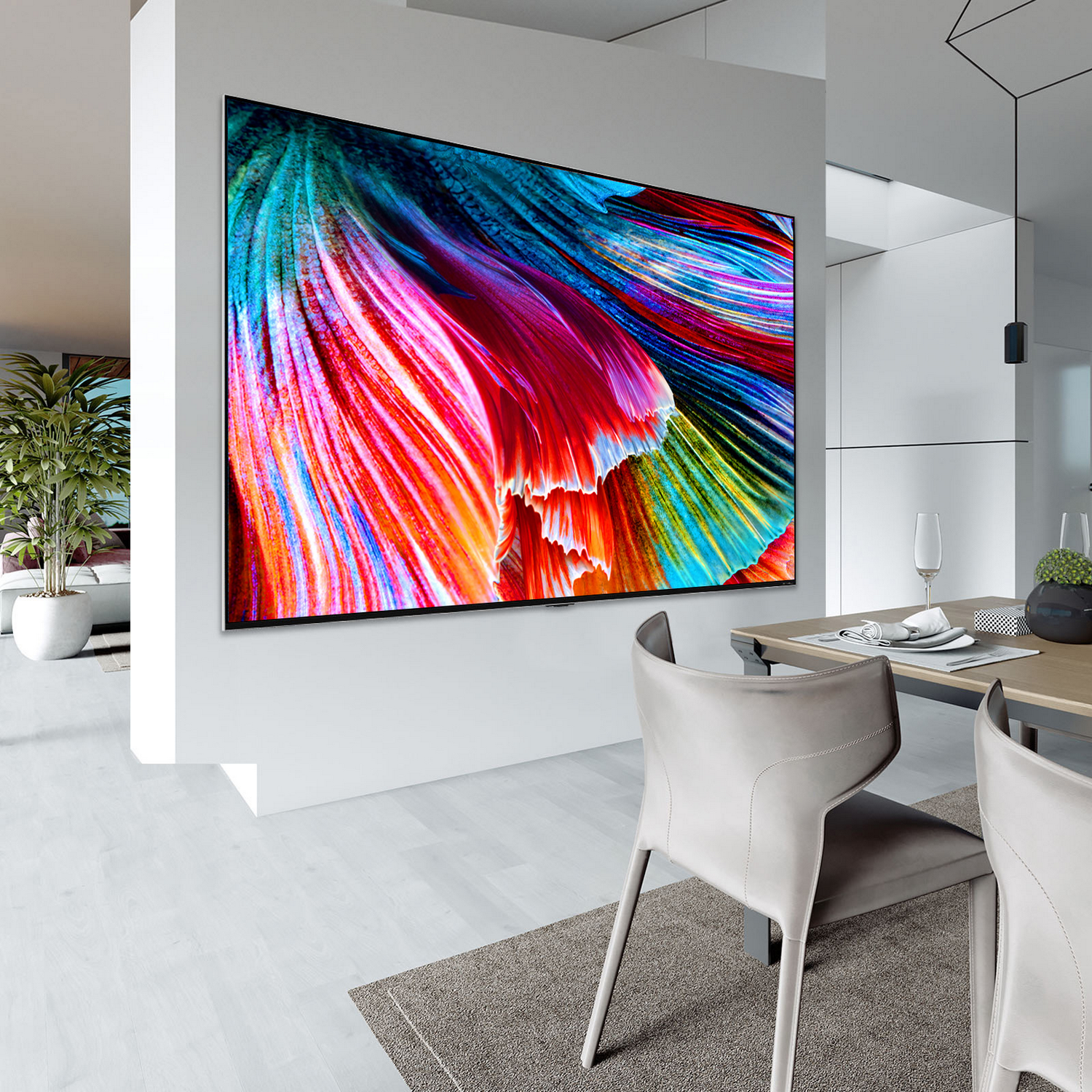Un televisor grande montado en una pared blanca. Muestra una imagen abstracta colorida. Debajo se ve un comedor.