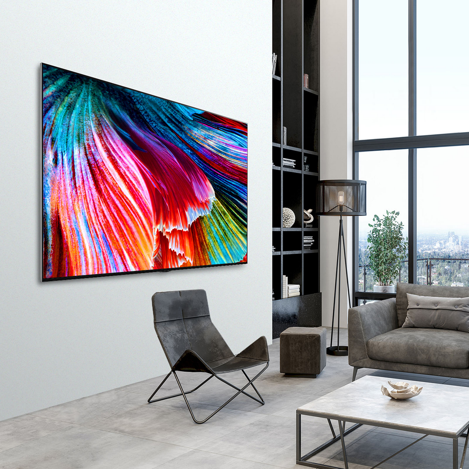 Un televisor grande muestra arte colorido. Una silla negra y un sofá gris están al frente. Una ventana muestra una vista de la ciudad.