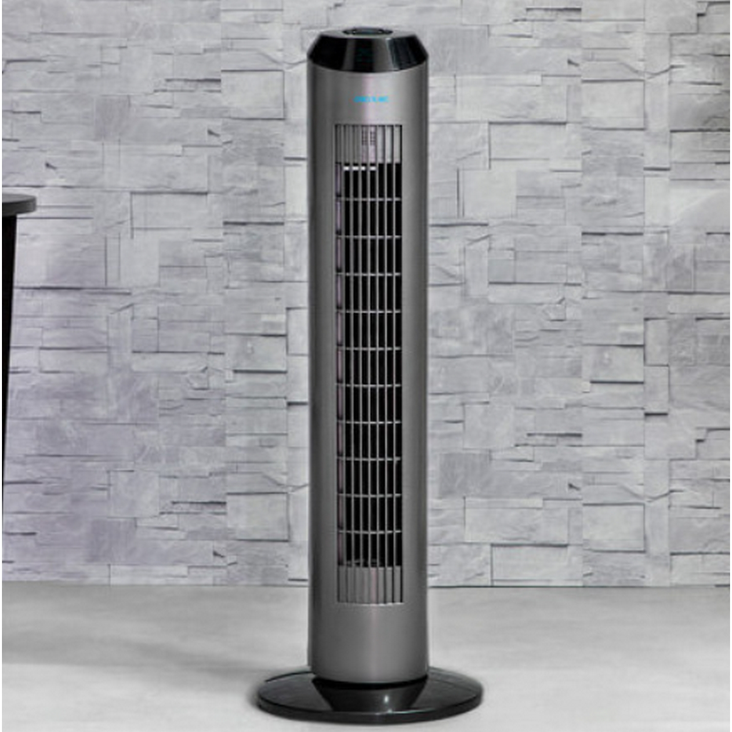 Un ventilador de torre cilíndrico gris sobre una base negra, contra una pared de ladrillo clara y texturizada.