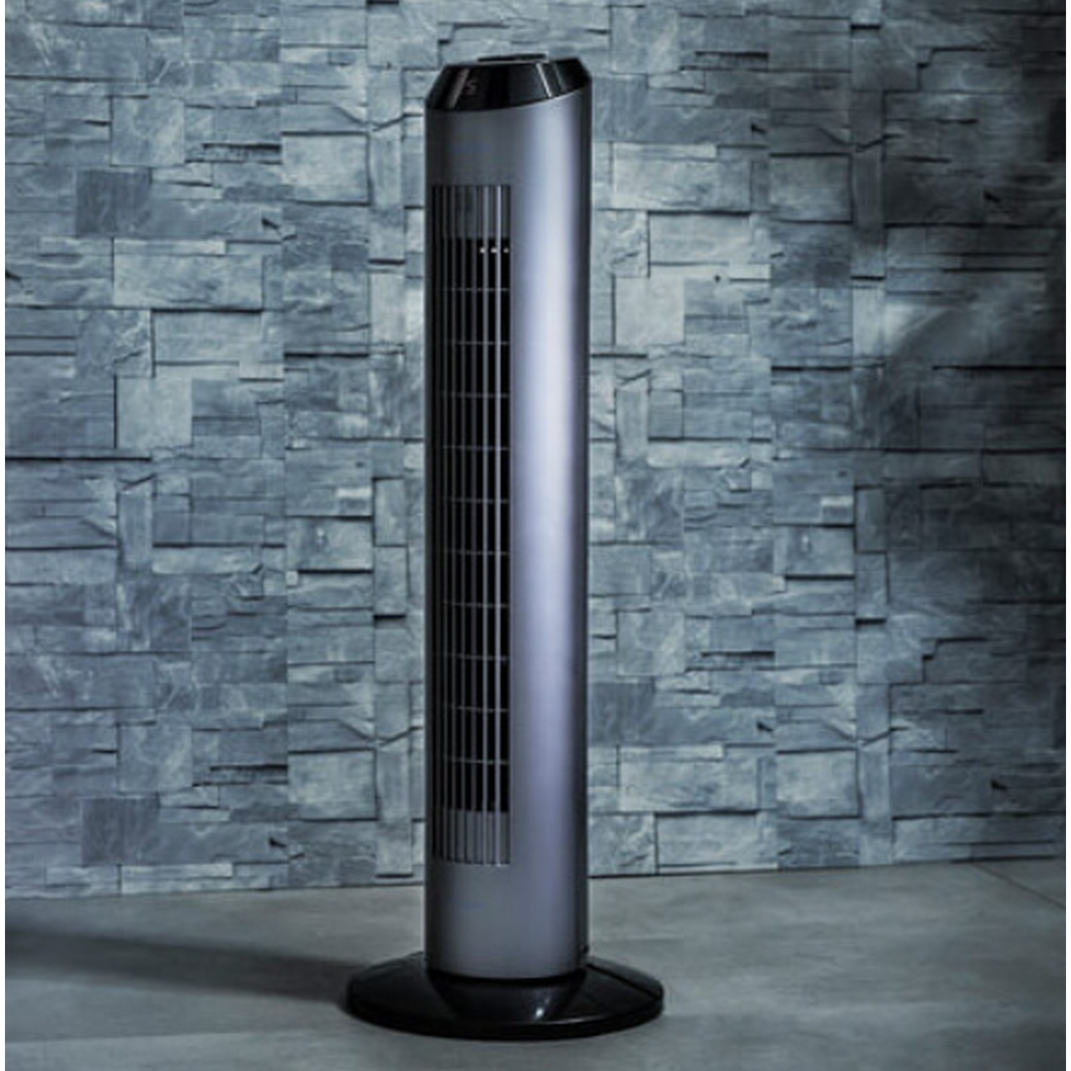 Ventilador de torre alto, delgado, plateado y negro, sobre una pared de bloques de piedra.