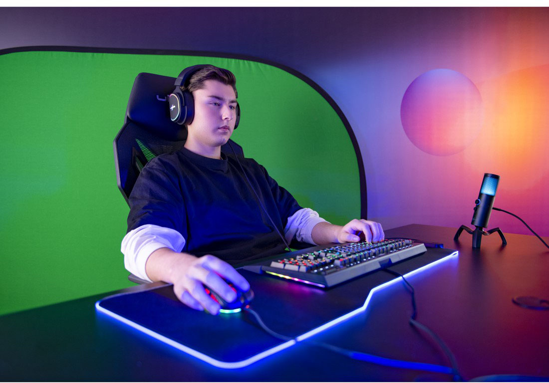 Gamer ül egy asztalnál, headsettel, billentyűzettel és mikrofonnal. Zöld háttér és egy lila gömb.