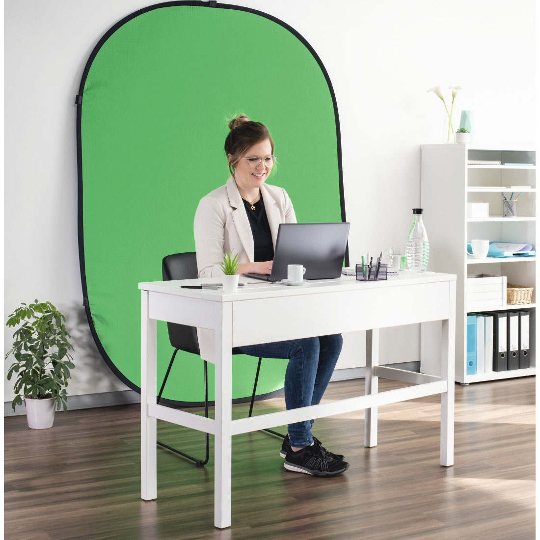 Nő egy fehér asztalnál, laptopon dolgozik. Zöld háttér. Fehér polcok jobbra.