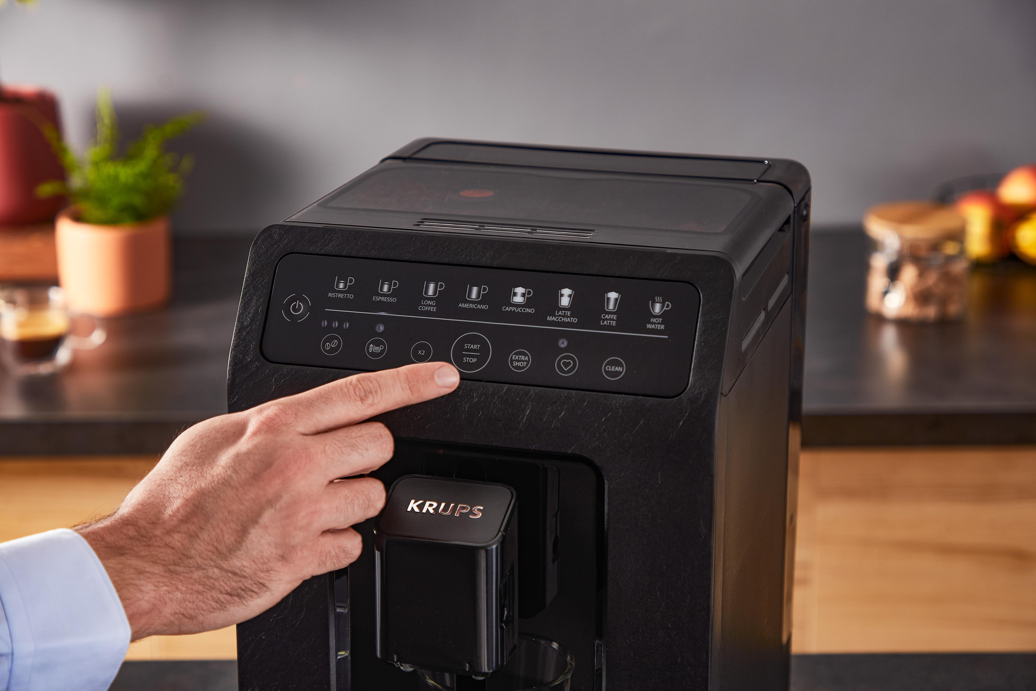 Eine Hand drückt eine Kaffeemaschinentaste. Tasten umfassen Espresso, Cappuccino und andere. Der Hintergrund zeigt eine Küche.