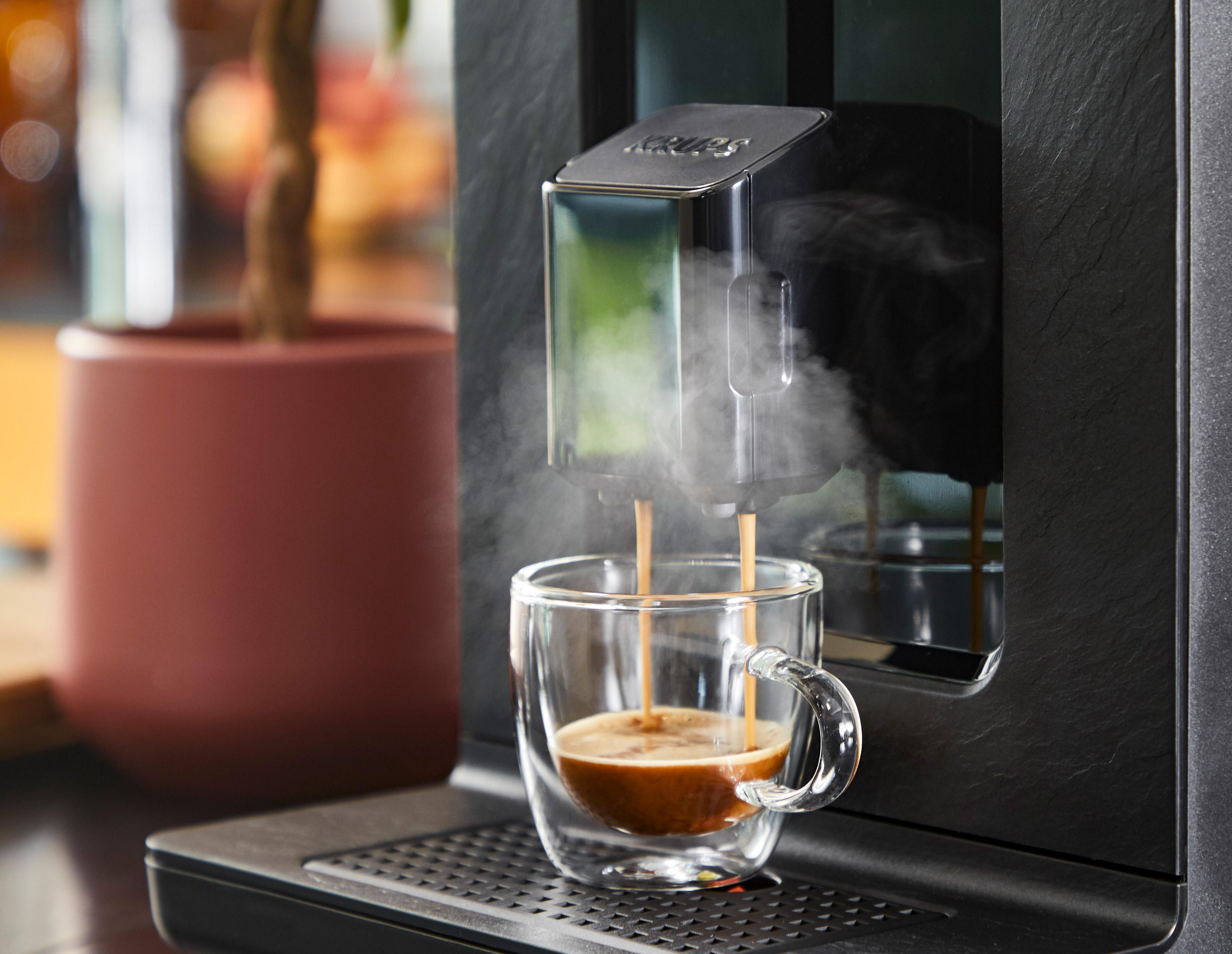 Espressomaschine, die Kaffee in eine doppelwandige Glastasse brüht; Dampf steigt auf, dunkle Farben.