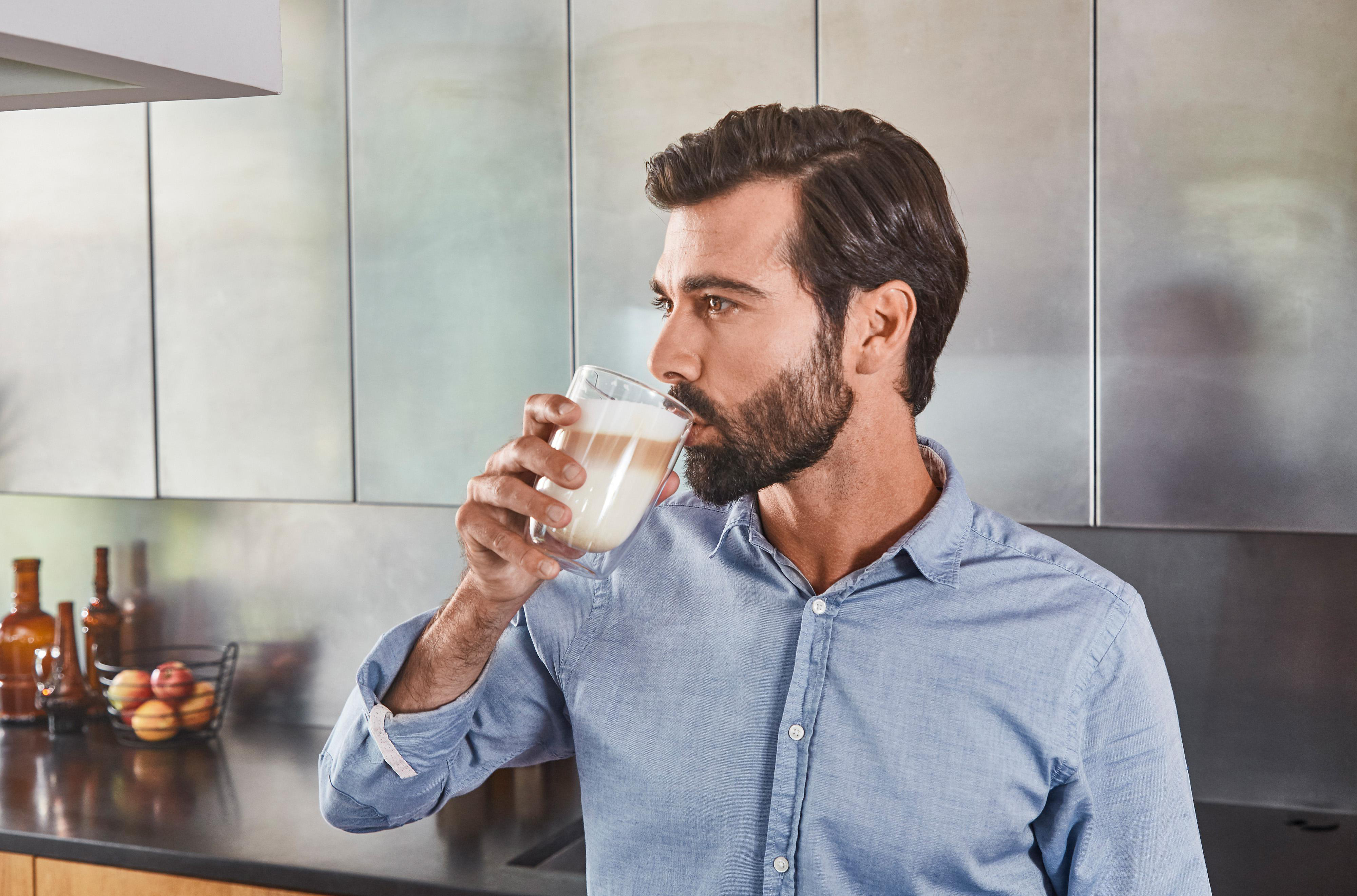Mann trinkt Latte Macchiato in moderner Küche, schaut zur Seite.