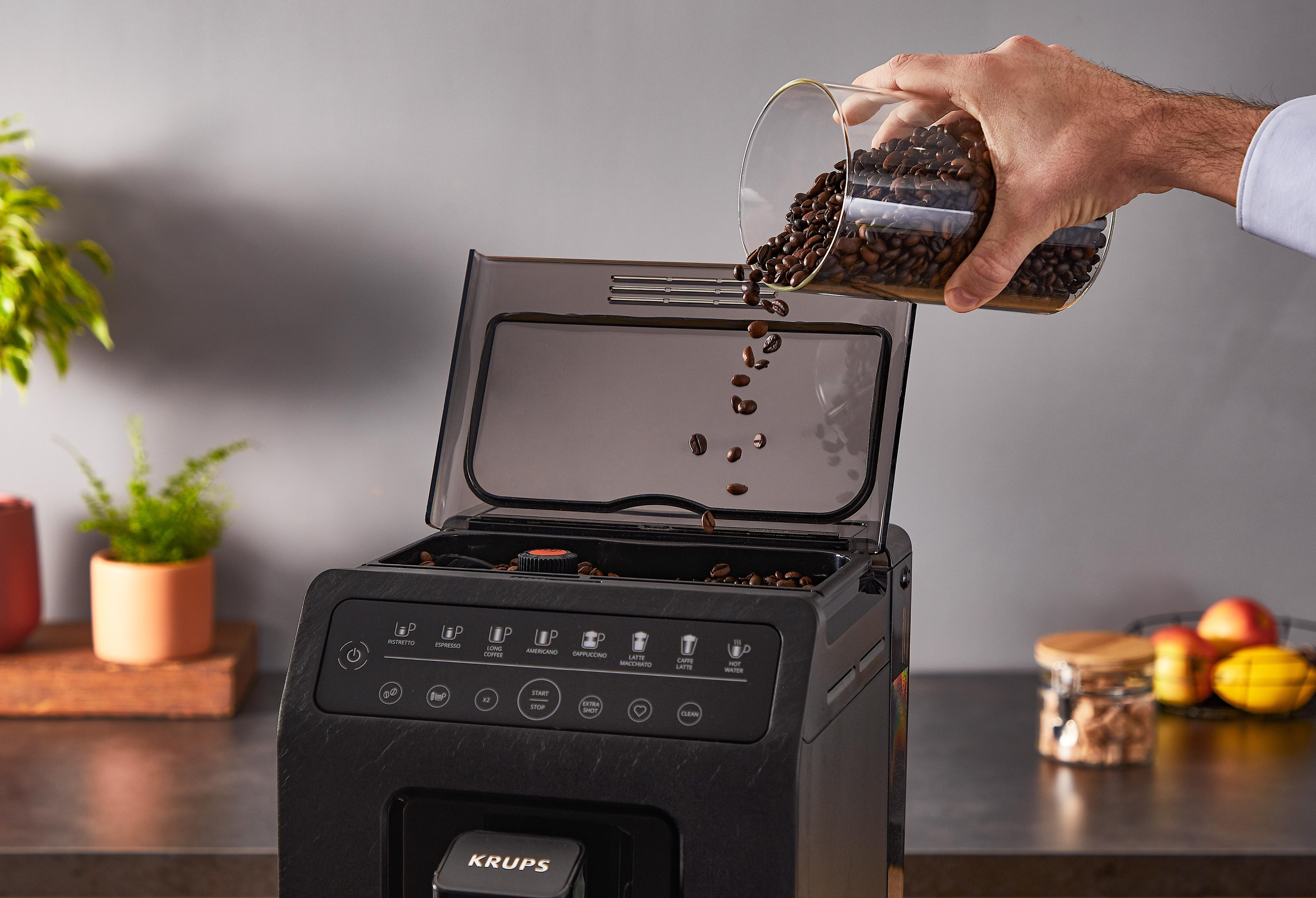 Eine Hand, die Kaffeebohnen in eine schwarze Kaffeemaschine gießt. Die Szene ist drinnen.