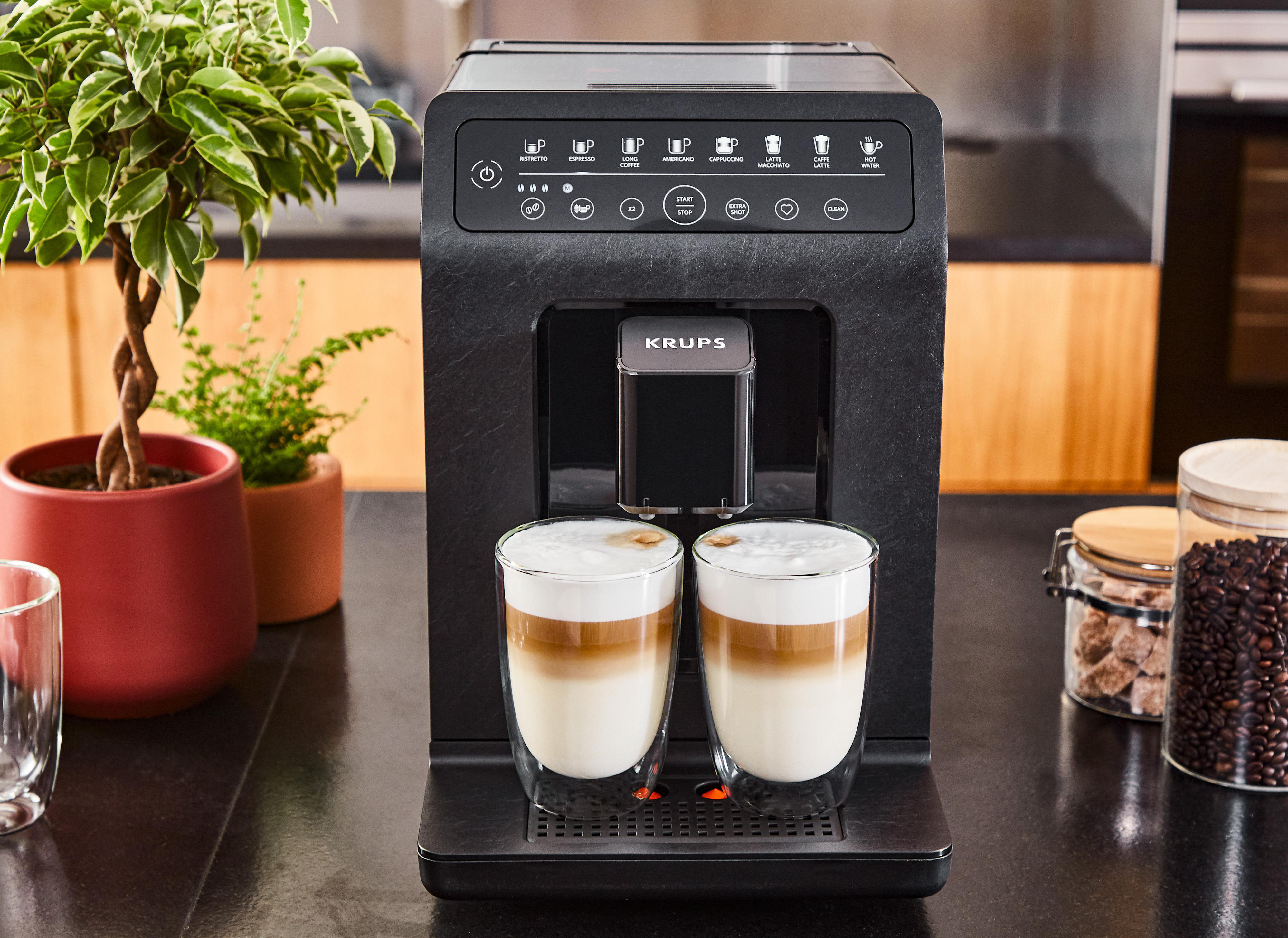 Eine schwarze Krups Kaffeemaschine mit zwei Tassen Latte, Bohnen und einem Zuckergas auf schwarzer Arbeitsplatte.