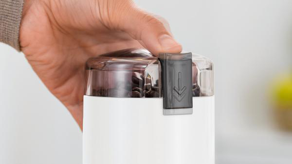 Hand bedient eine weiße Kaffeemühle mit transparentem Deckel, der Kaffeebohnen zeigt.