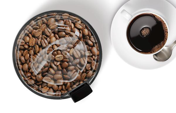 Eine Kaffeemühle gefüllt mit Kaffeebohnen, neben einer Tasse Kaffee auf weißem Hintergrund.