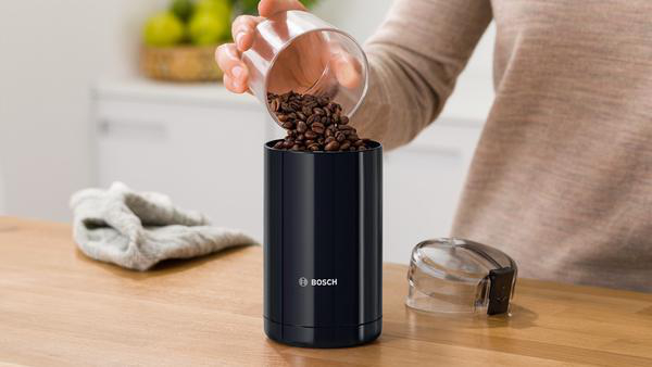 Eine Person gießt Kaffeebohnen in eine schwarze Kaffeemühle. Ein Tuch und ein Deckel sind ebenfalls sichtbar.