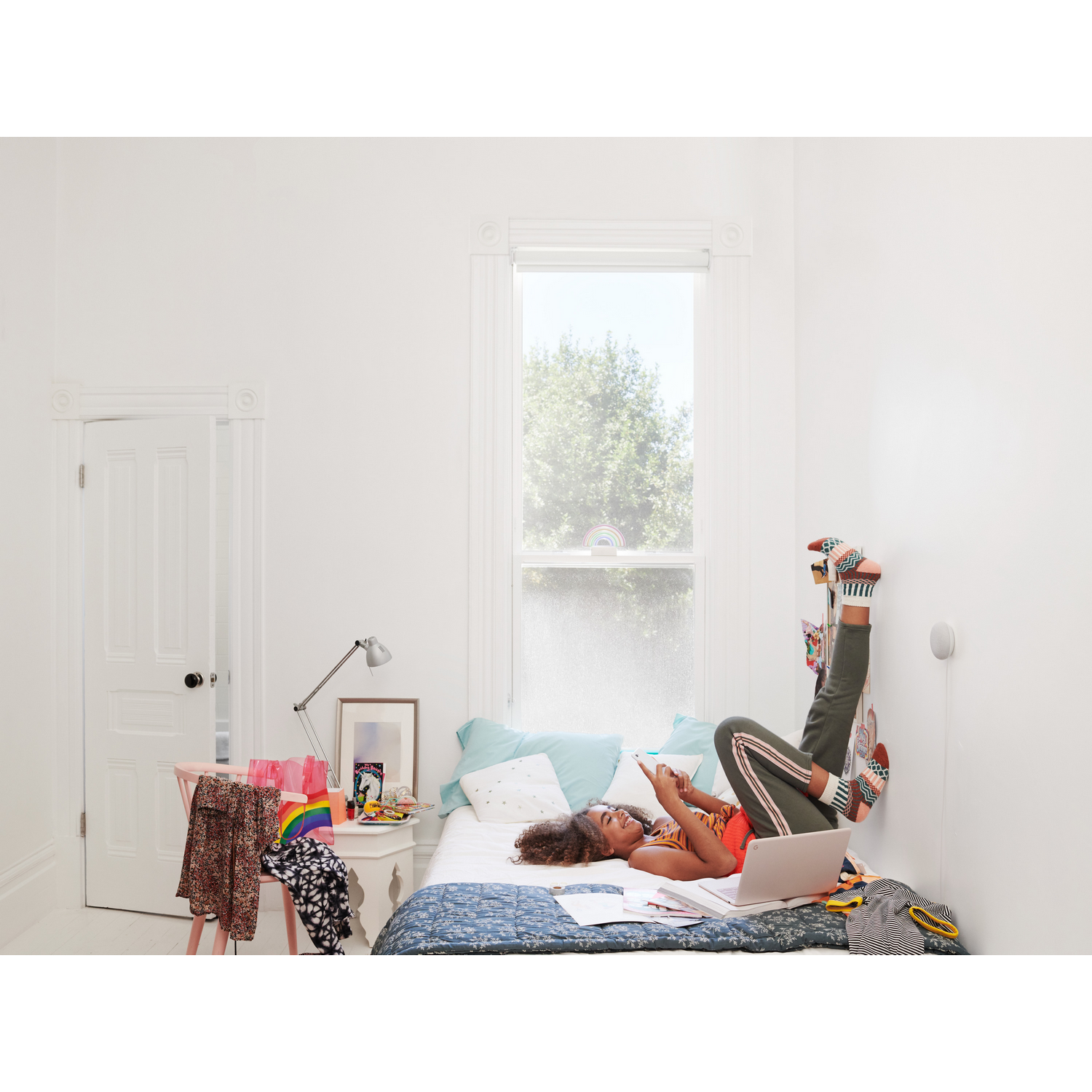 Una mujer acostada en una cama usa un teléfono. La habitación es blanca con una ventana y un escritorio pequeño con ropa.
