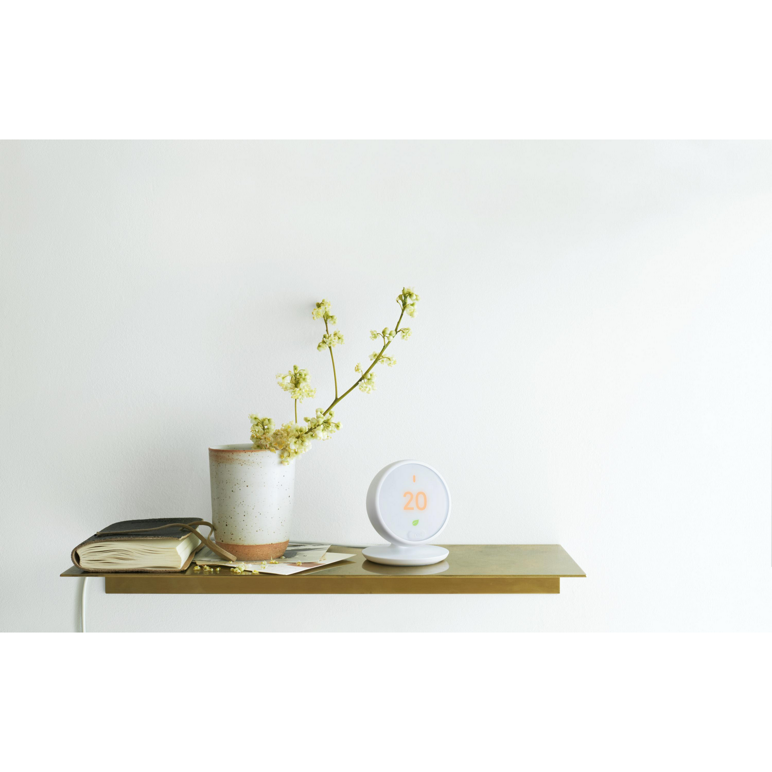 Un termostato inteligente moderno y un libro con un florero en un estante dorado contra una pared blanca.