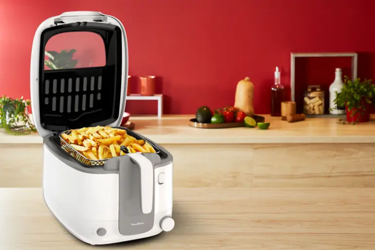 Een wit en grijze friteuse met friet, rode muur en keukenartikelen.